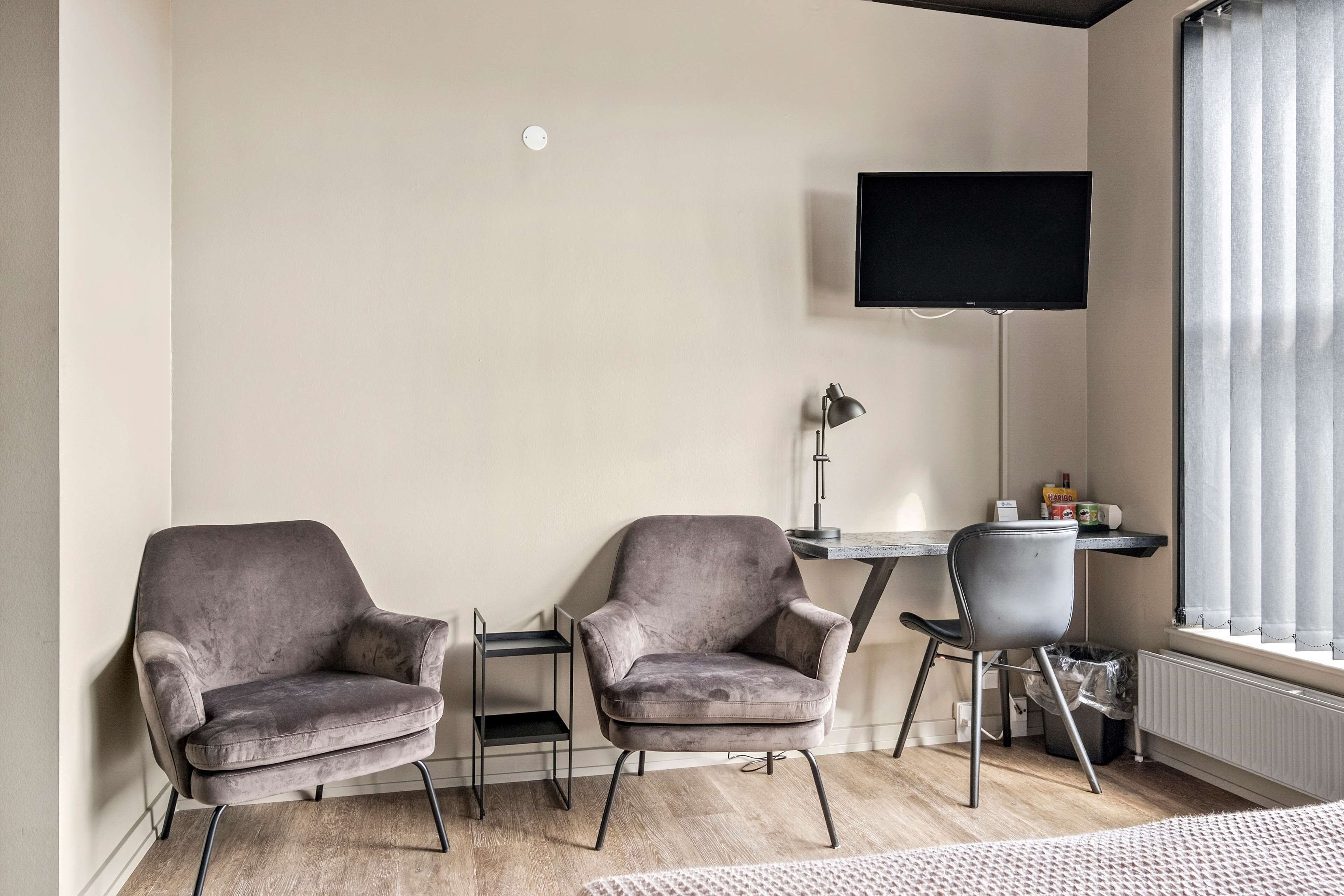 A modern living room with two grey armchairs, a small table, a TV mounted on the wall, and a window with curtains.