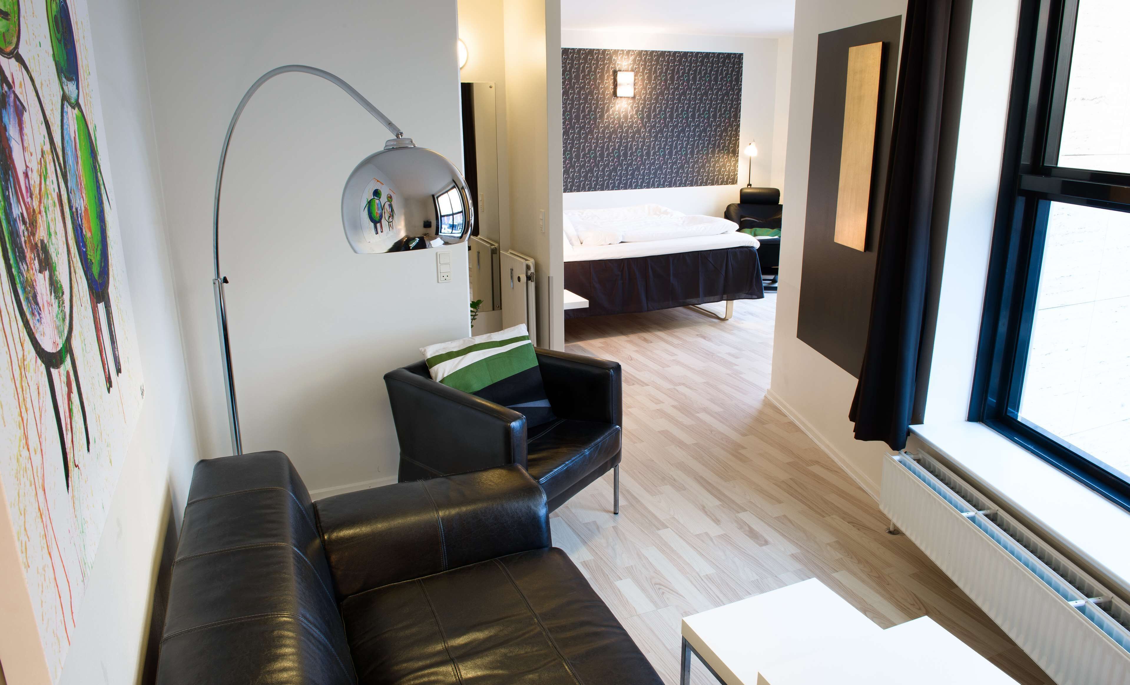 A modern hotel room with a black leather sofa, a small white table, a large window with blinds, a bed with a dark headboard, and a wall-mounted desk lamp.