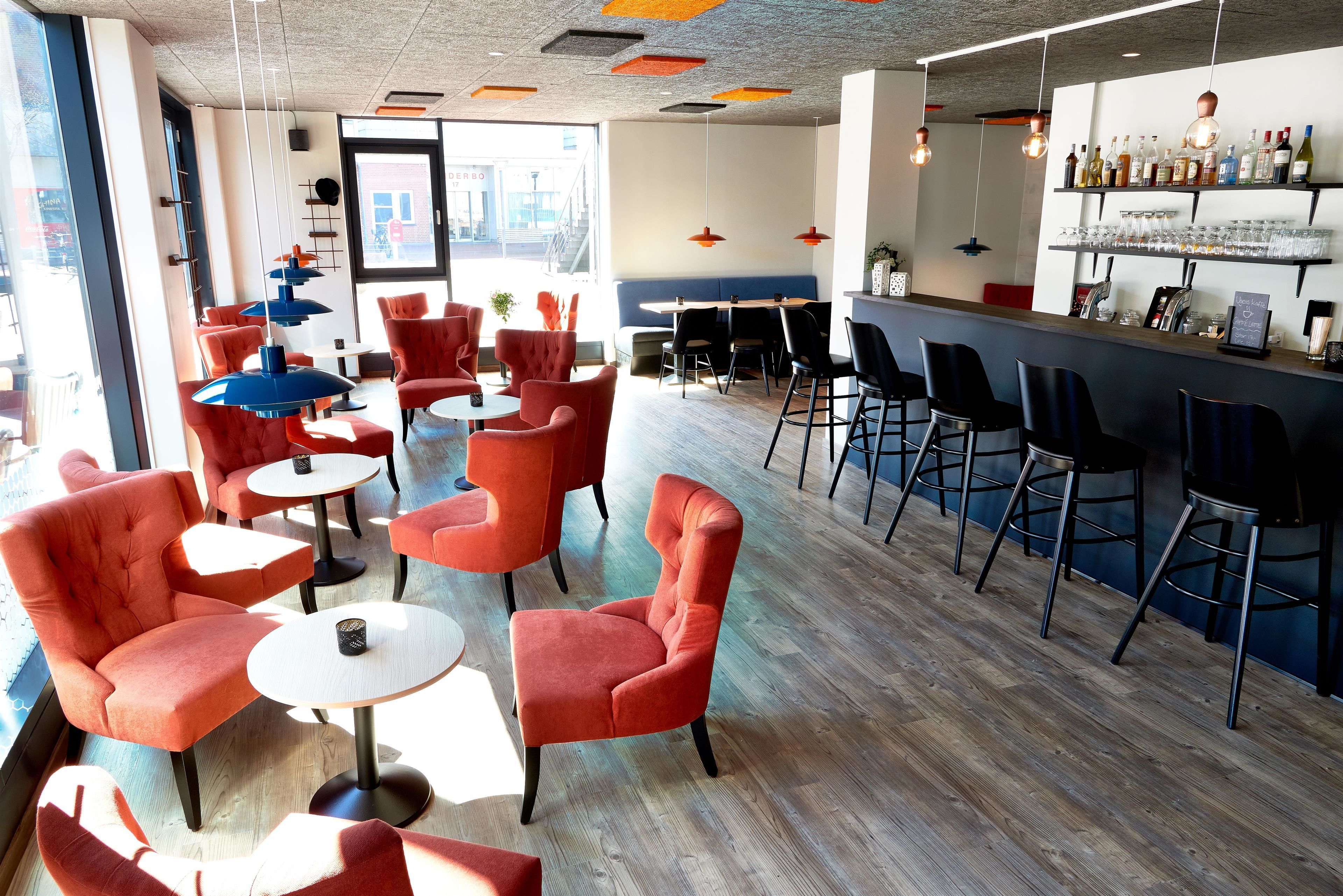 A modern bar area with red and black bar stools, a variety of bottles and glasses on shelves, and a seating area with red chairs and round tables.