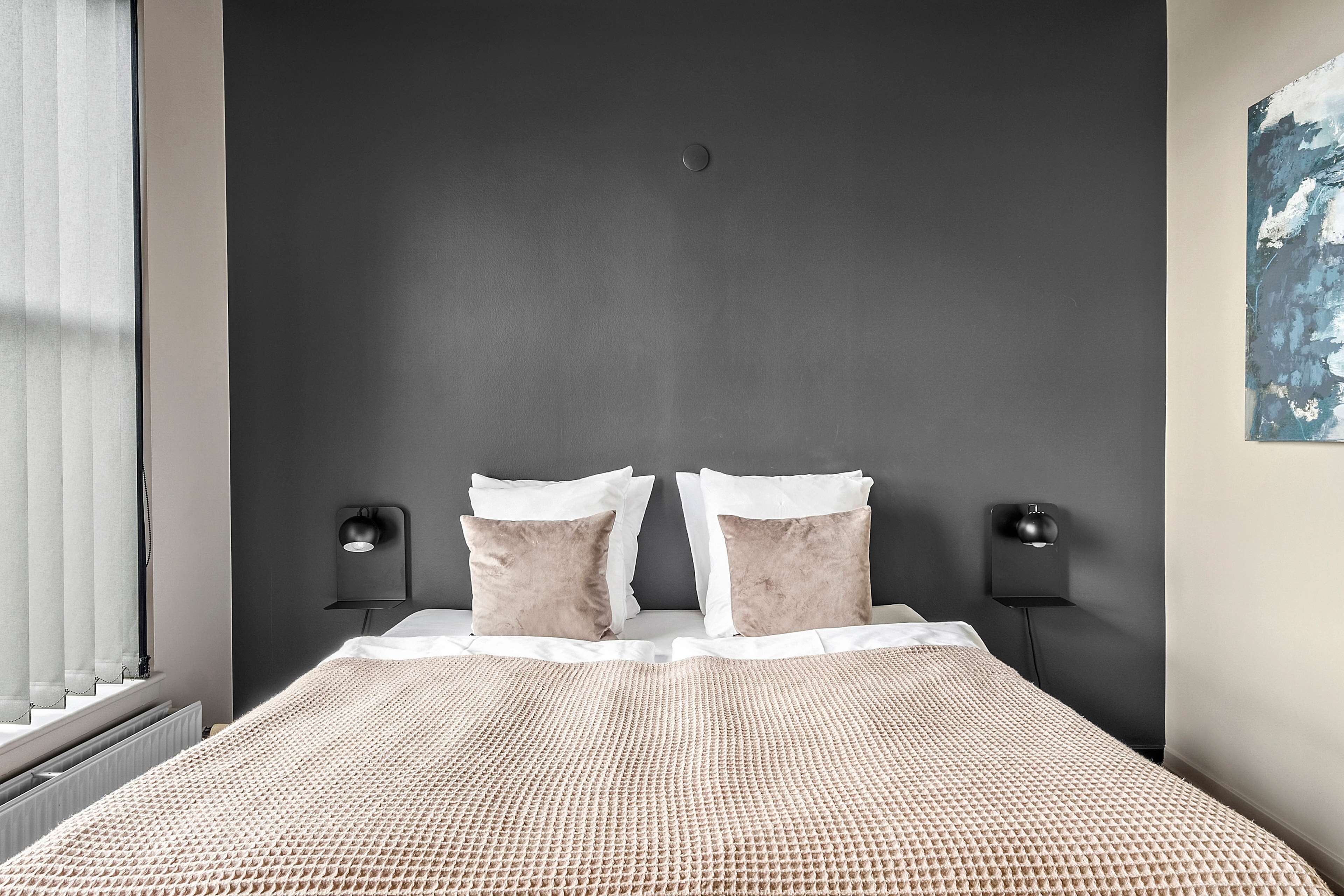 Un dormitorio moderno con una pared oscura, dos lámparas de noche, una cama con colcha beige estampada y un ventanal con cortinas traslúcidas.