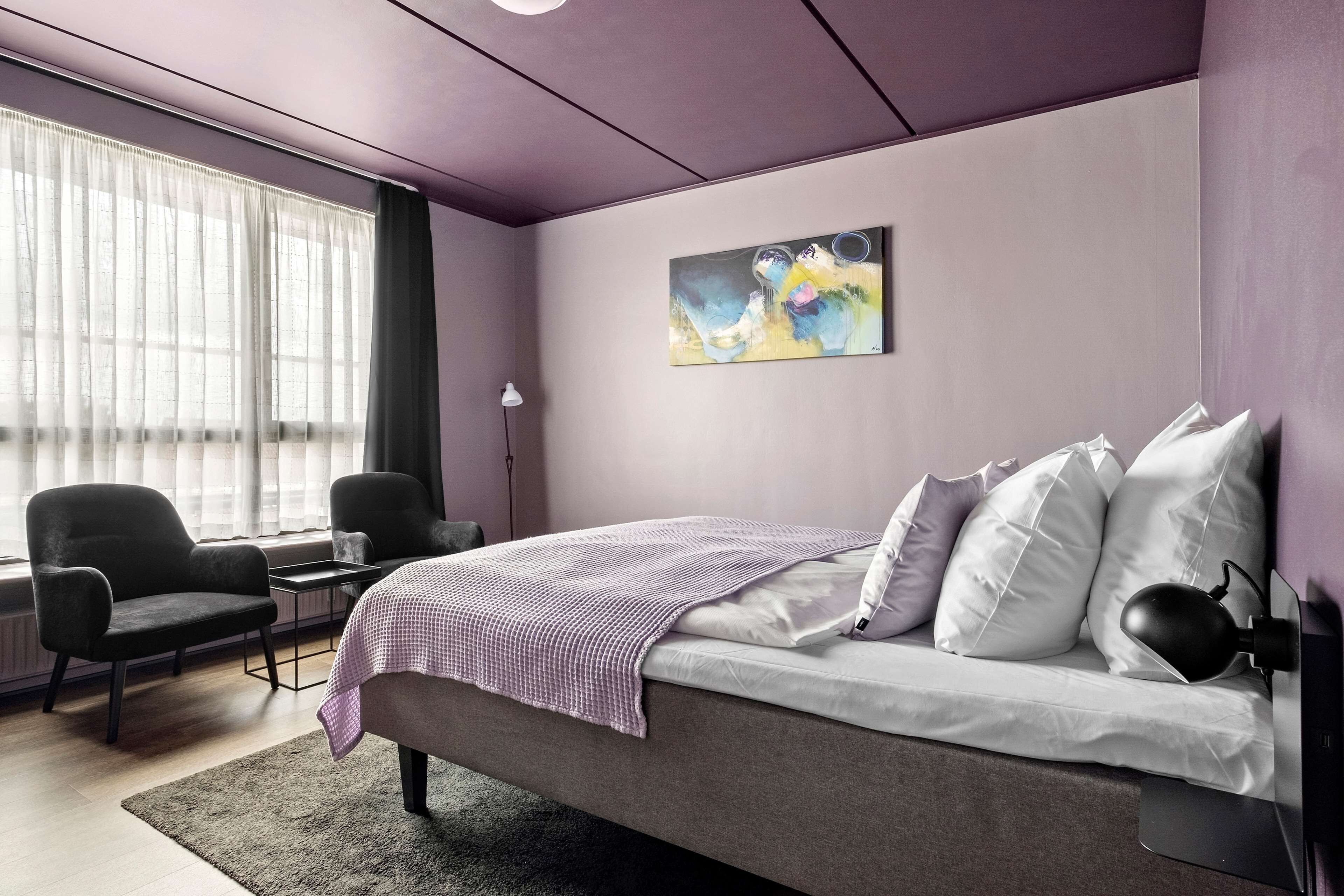 A modern bedroom with a bed, two chairs, a desk, and a large window with sheer curtains.