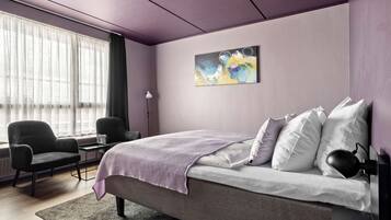 A modern bedroom with a bed, two chairs, a desk, and a large window with sheer curtains.