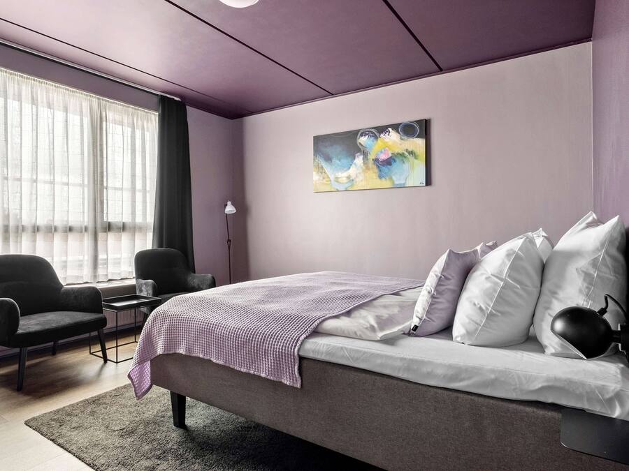 A modern bedroom with a bed, two chairs, a desk, and a large window with sheer curtains.