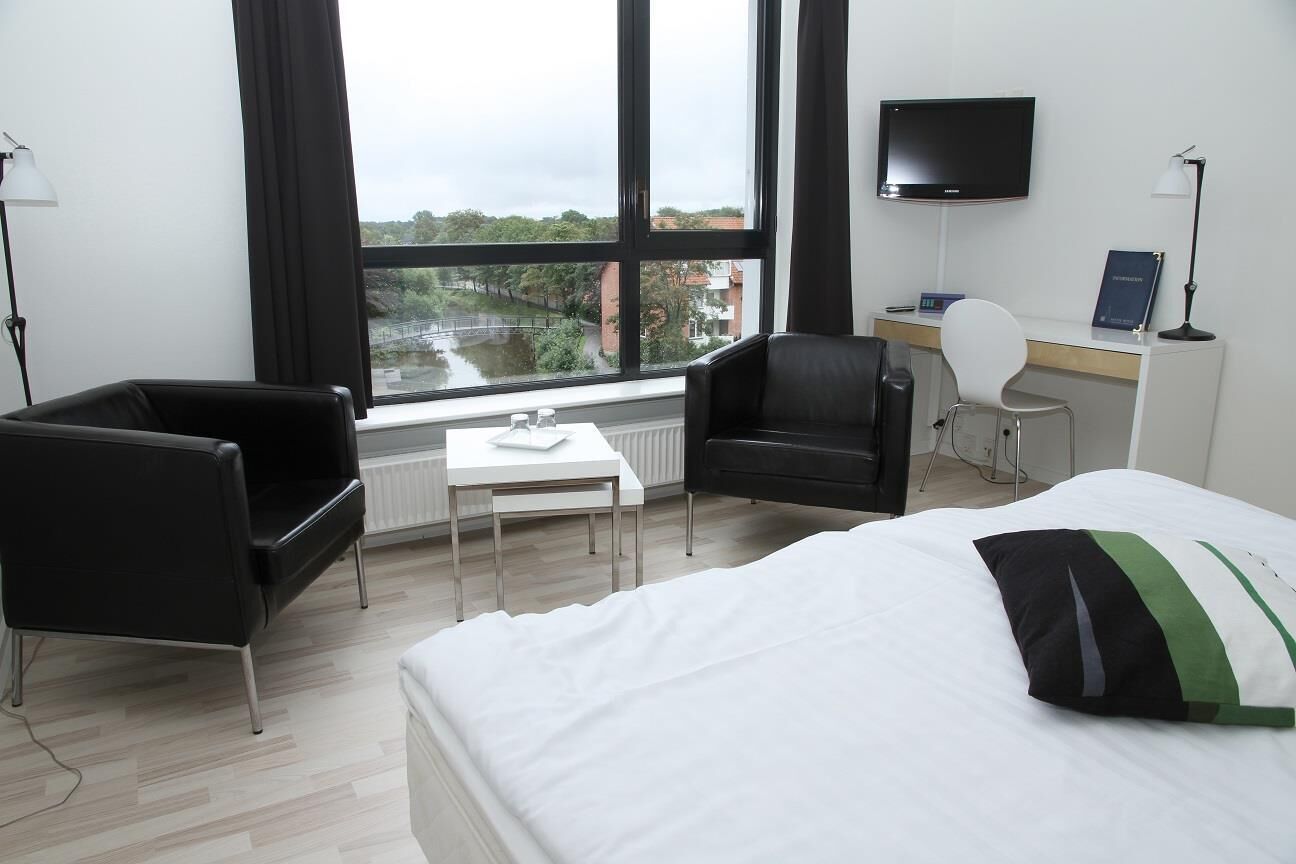 Une chambre d’hôtel équipée d’un lit, d’un bureau, d’une chaise, d’une télévision et d’une fenêtre donnant sur une rivière et des bâtiments.