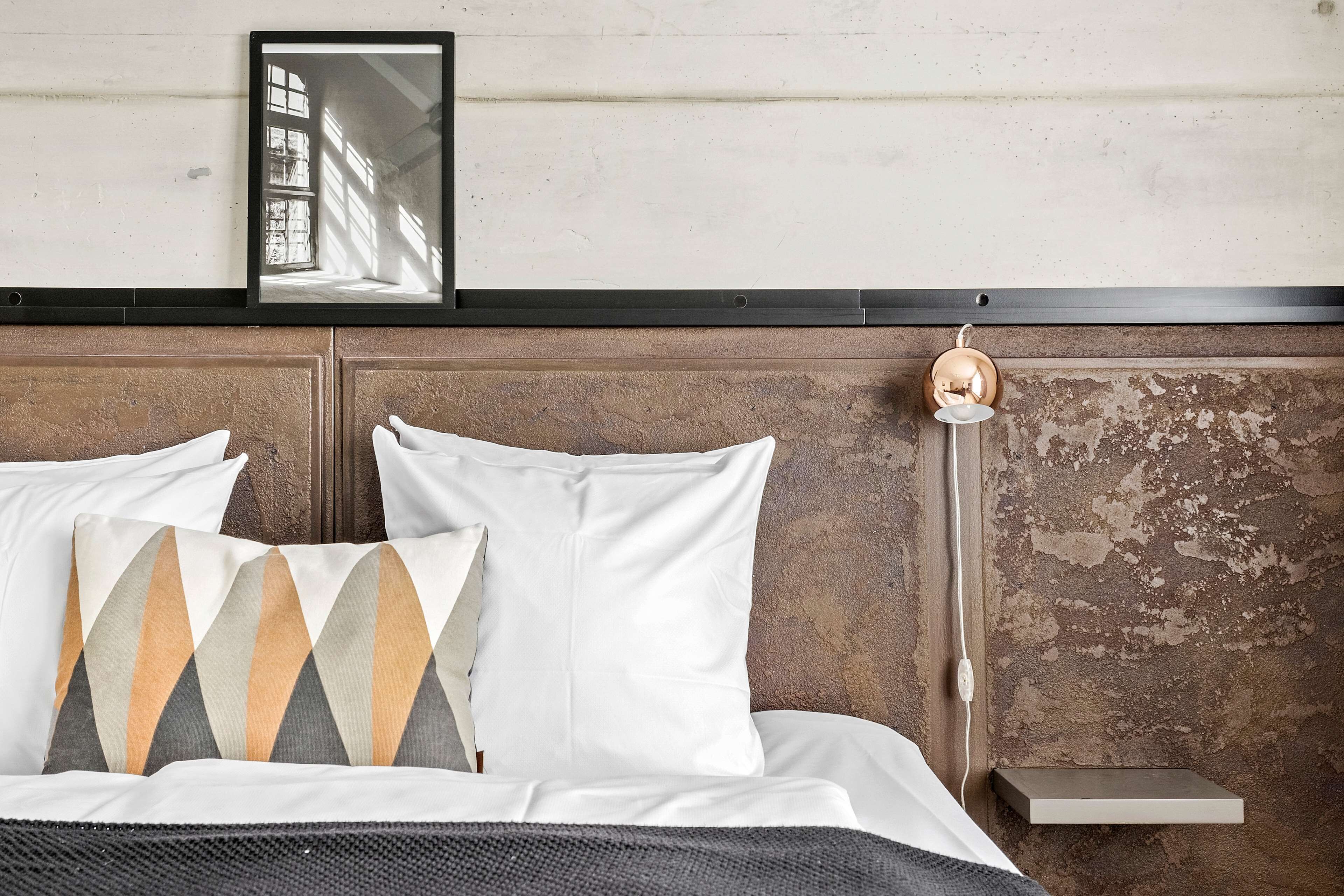 A bed with white bedding and patterned pillows, a bedside table with a lamp, and a wall-mounted shelf.