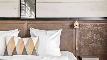 A bed with white bedding and patterned pillows, a bedside table with a lamp, and a wall-mounted shelf.
