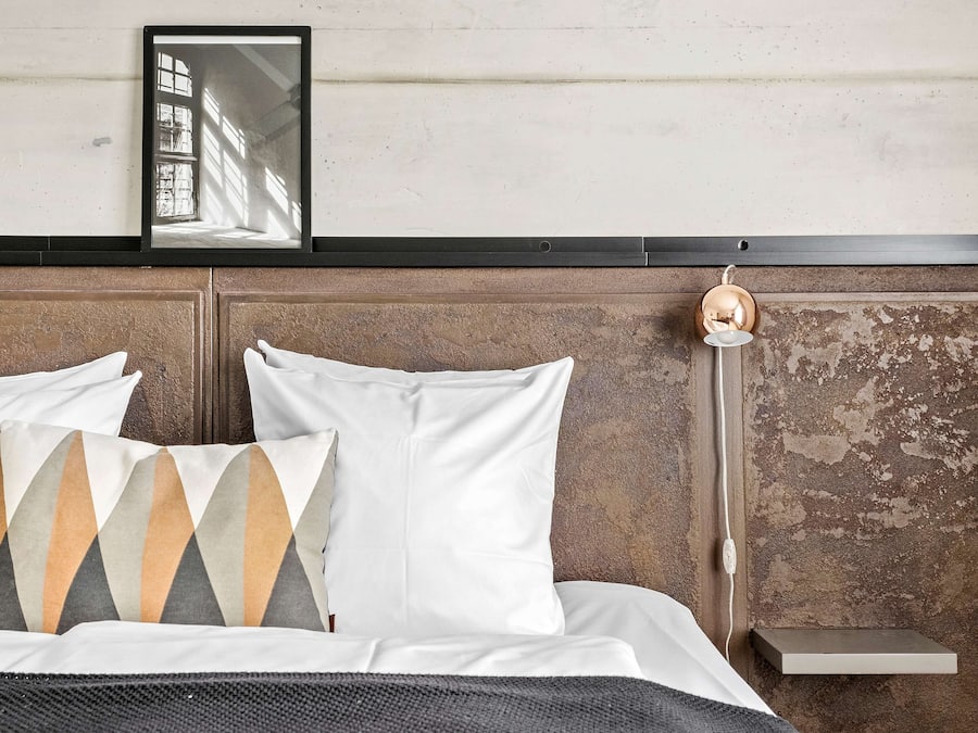 A bed with white bedding and patterned pillows, a bedside table with a lamp, and a wall-mounted shelf.
