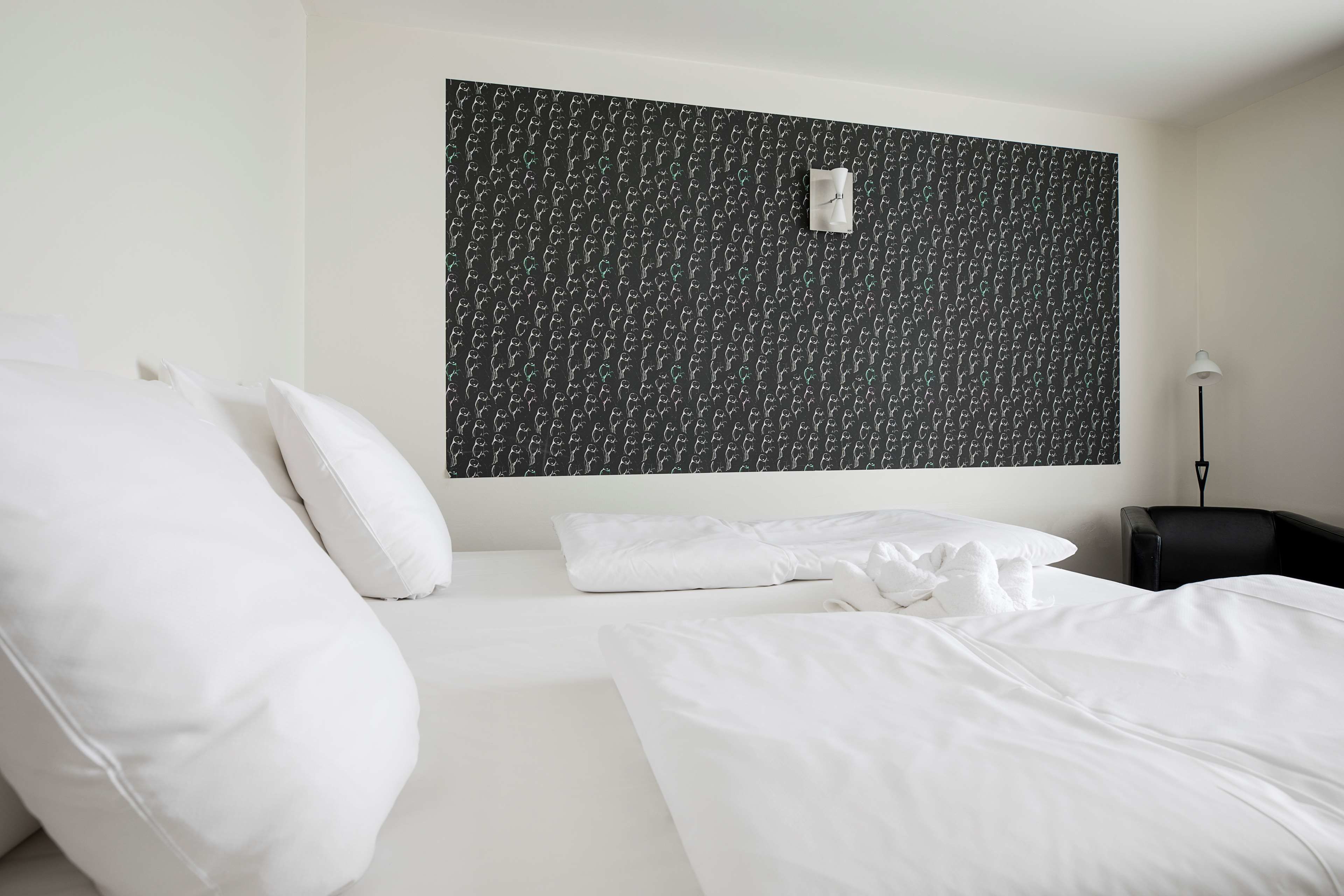 A hotel room with a neatly made bed, white bedding, a black patterned wall, and a small desk with a chair.