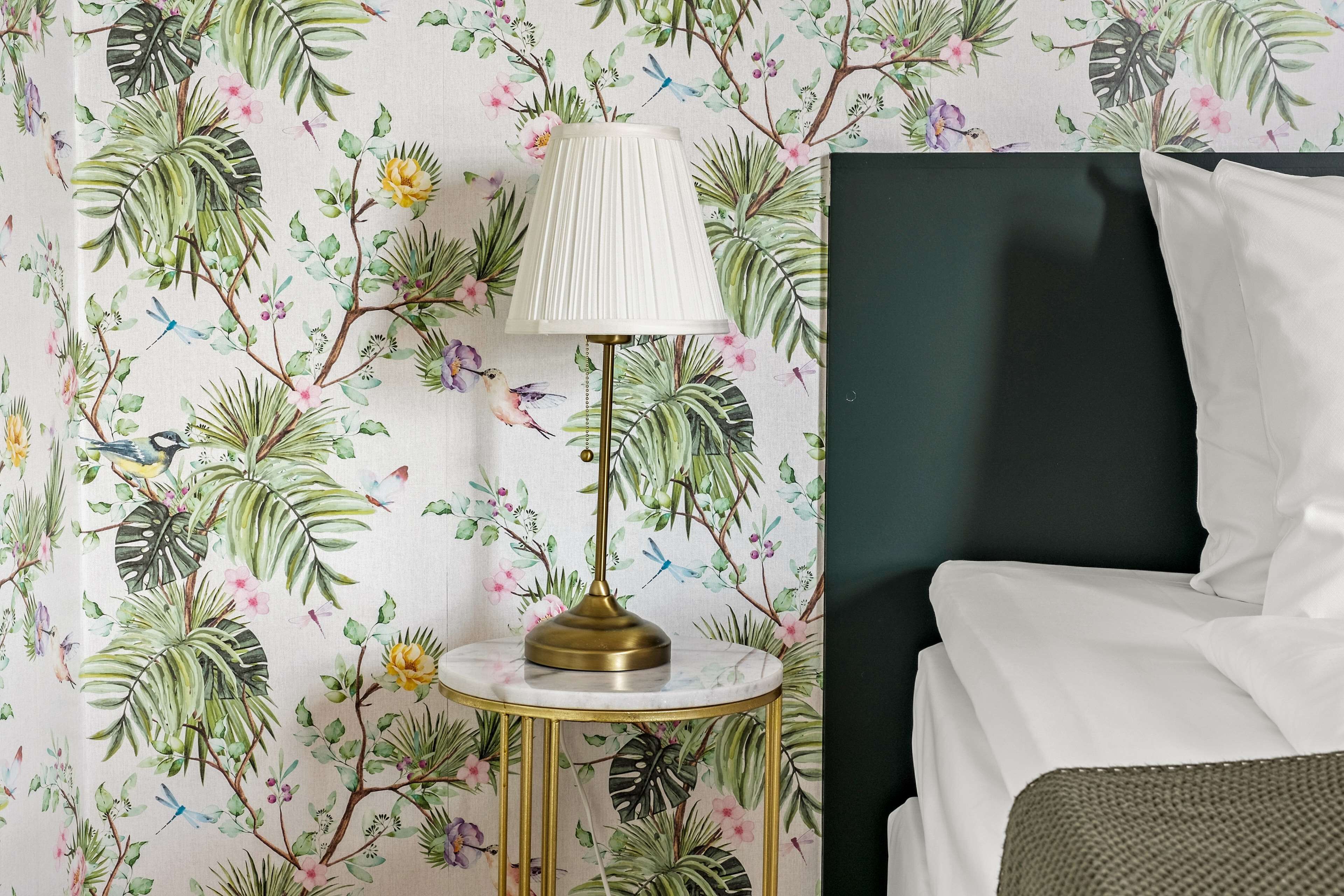 Una habitación con una cama, una mesa redonda con una lámpara y un papel tapiz con motivos florales.