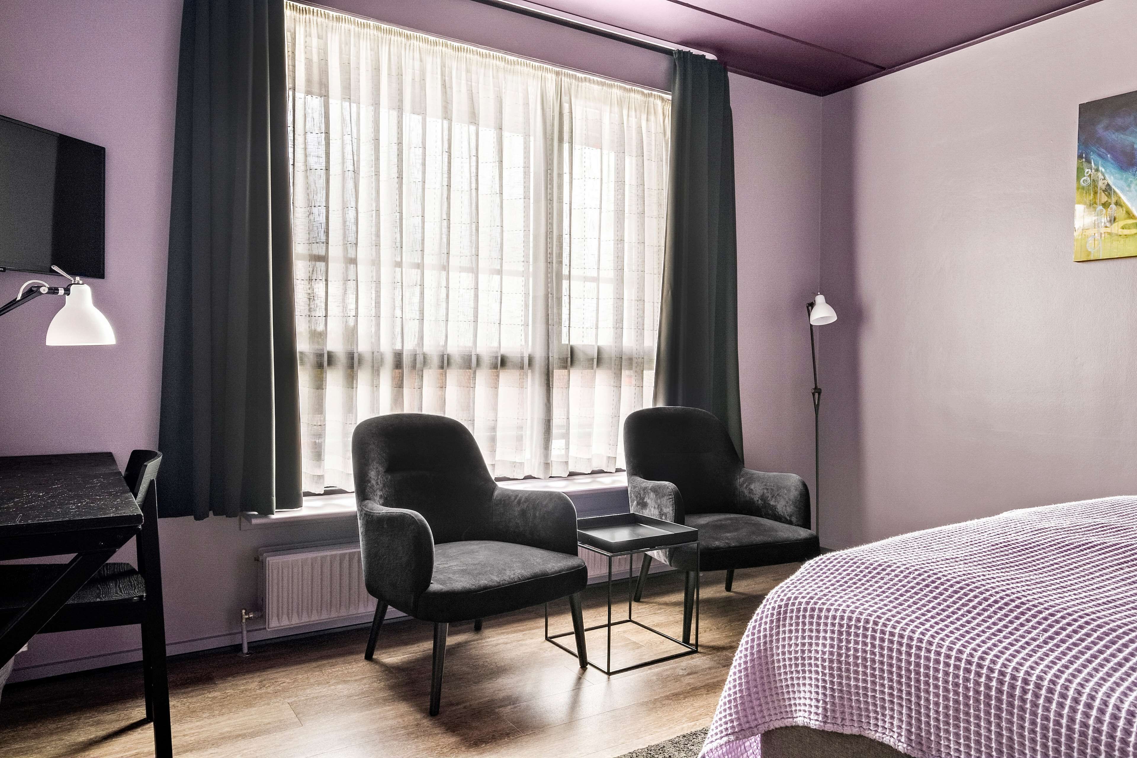 A room with a bed, two armchairs, a small table, a desk, a radiator, and a window with curtains.
