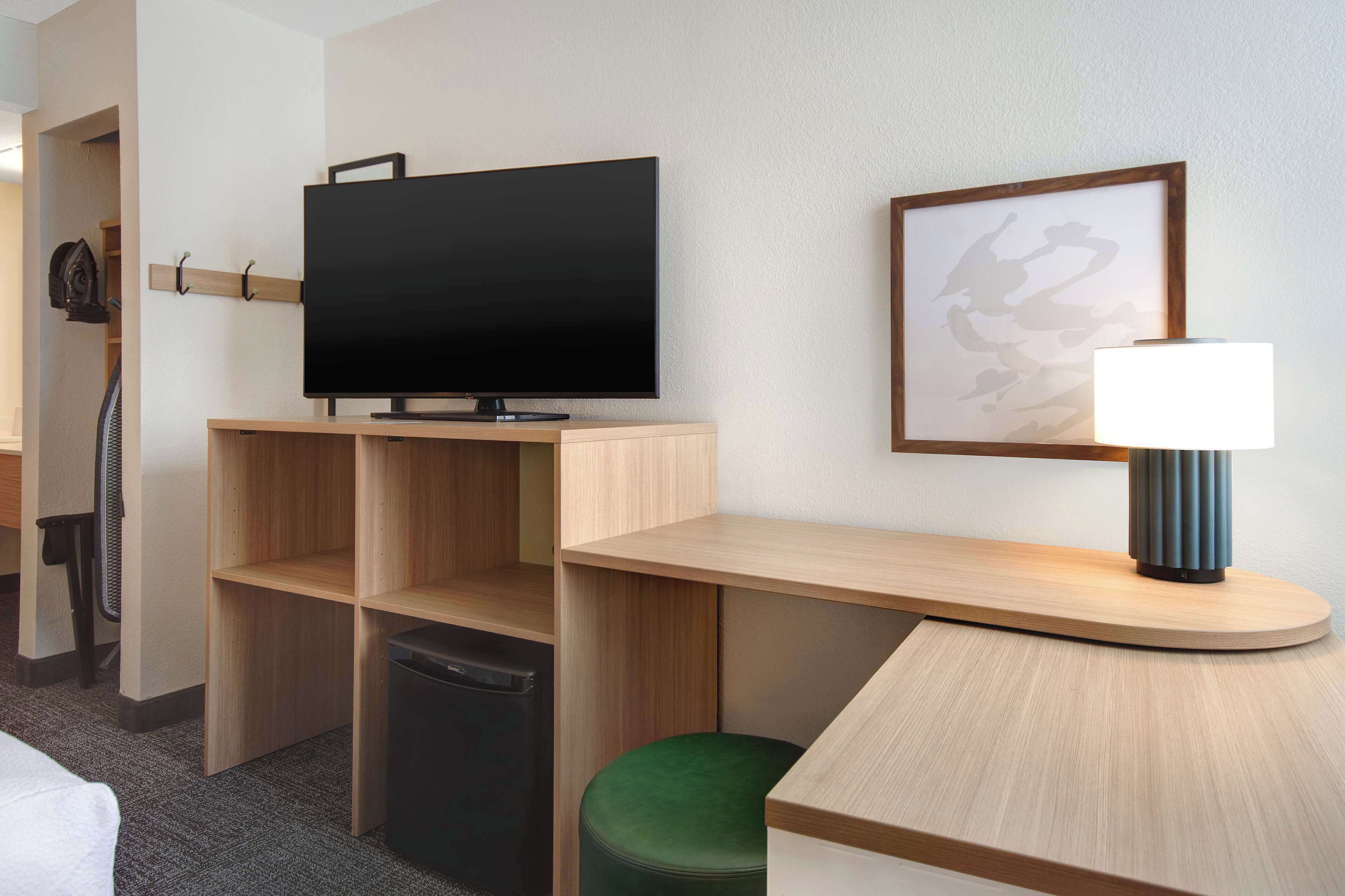 A hotel room with a flat-screen TV on a wooden stand, a lamp on a curved wooden table, a green ottoman, and a framed picture on the wall.