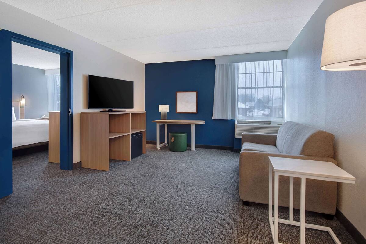 A hotel room with a bed, a TV, a desk, a lamp, a green trash bin, and a window with curtains.