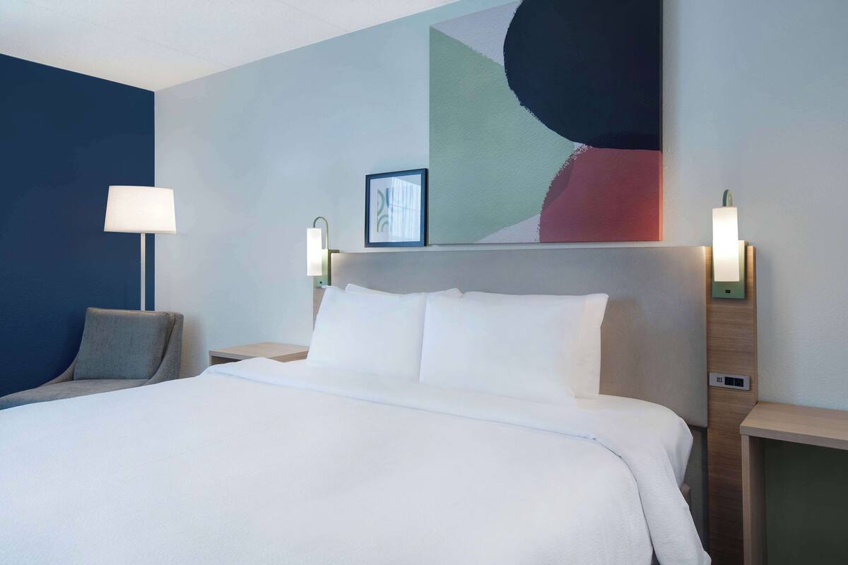 A modern hotel room with a large bed, two bedside tables, a lamp, and a wall art piece.