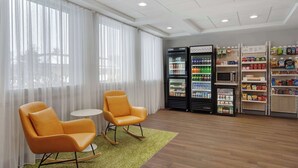 A hotel lobby with two orange chairs, a small table, a refrigerator stocked with beverages, and shelves with snacks. - Spark by Hilton Rochester University Area (Rochester)