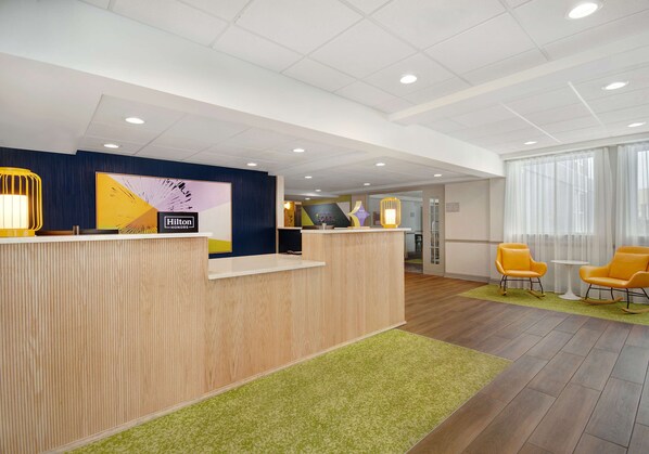 A hotel reception area with a wooden reception desk, a large digital screen displaying the Hilton logo, and a seating area with yellow chairs.