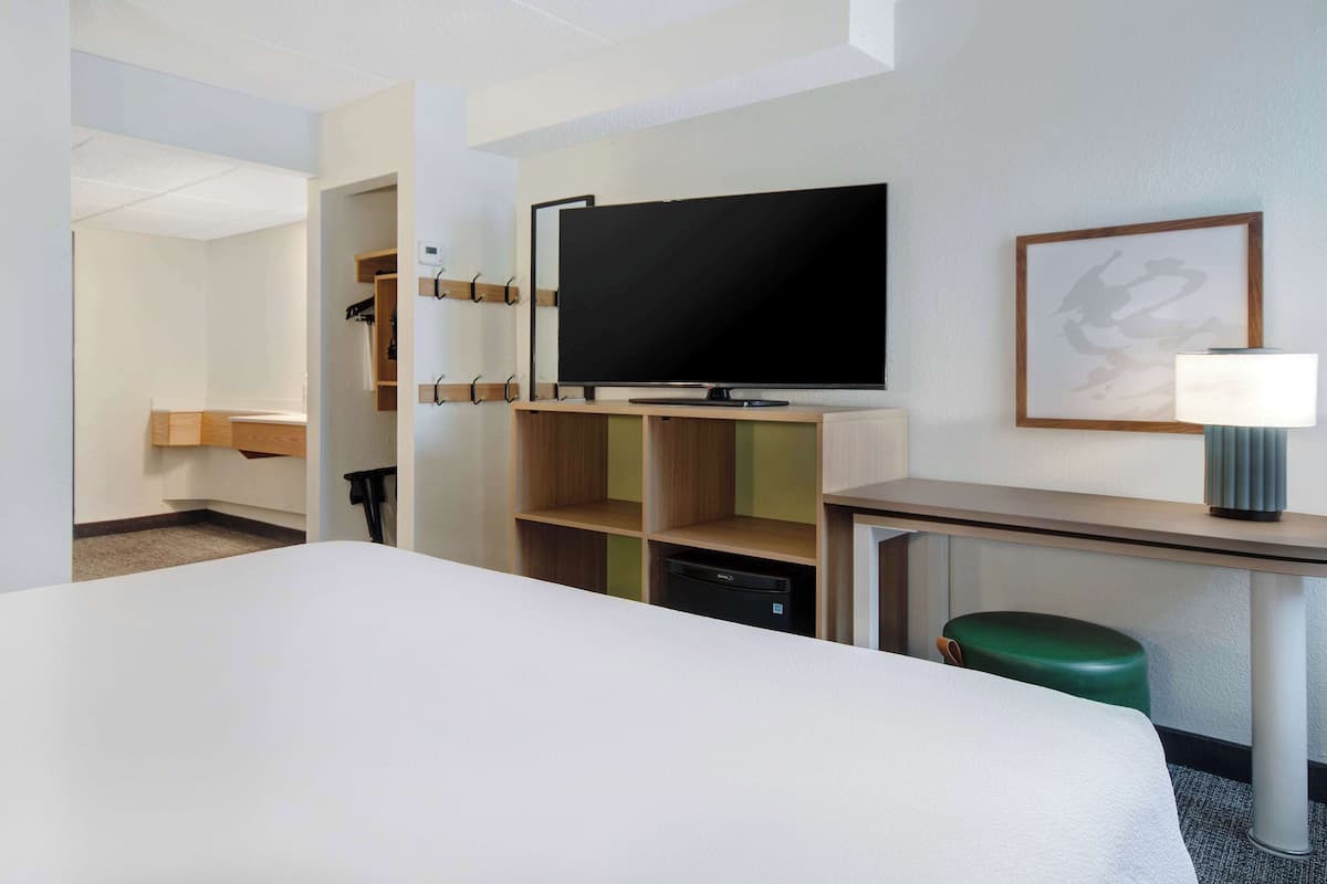 A hotel room with a bed, a desk with a television, a green stool, and a wall-mounted shelf.