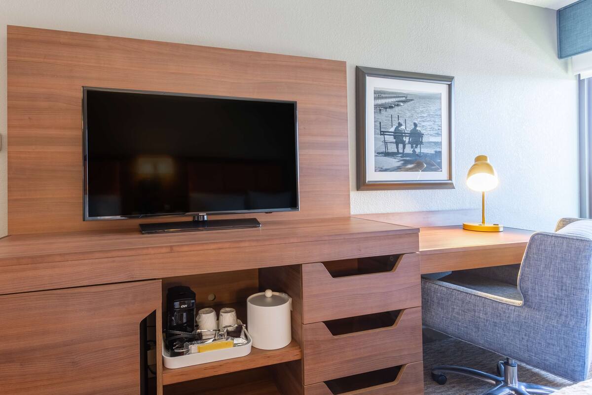 A hotel room with a wooden desk, a flat-screen TV, a framed picture on the wall, and a desk lamp.
