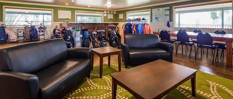 A room with two black leather sofas, a wooden coffee table, and a green carpet with a white pattern. There are golf bags and clothing racks in the background.