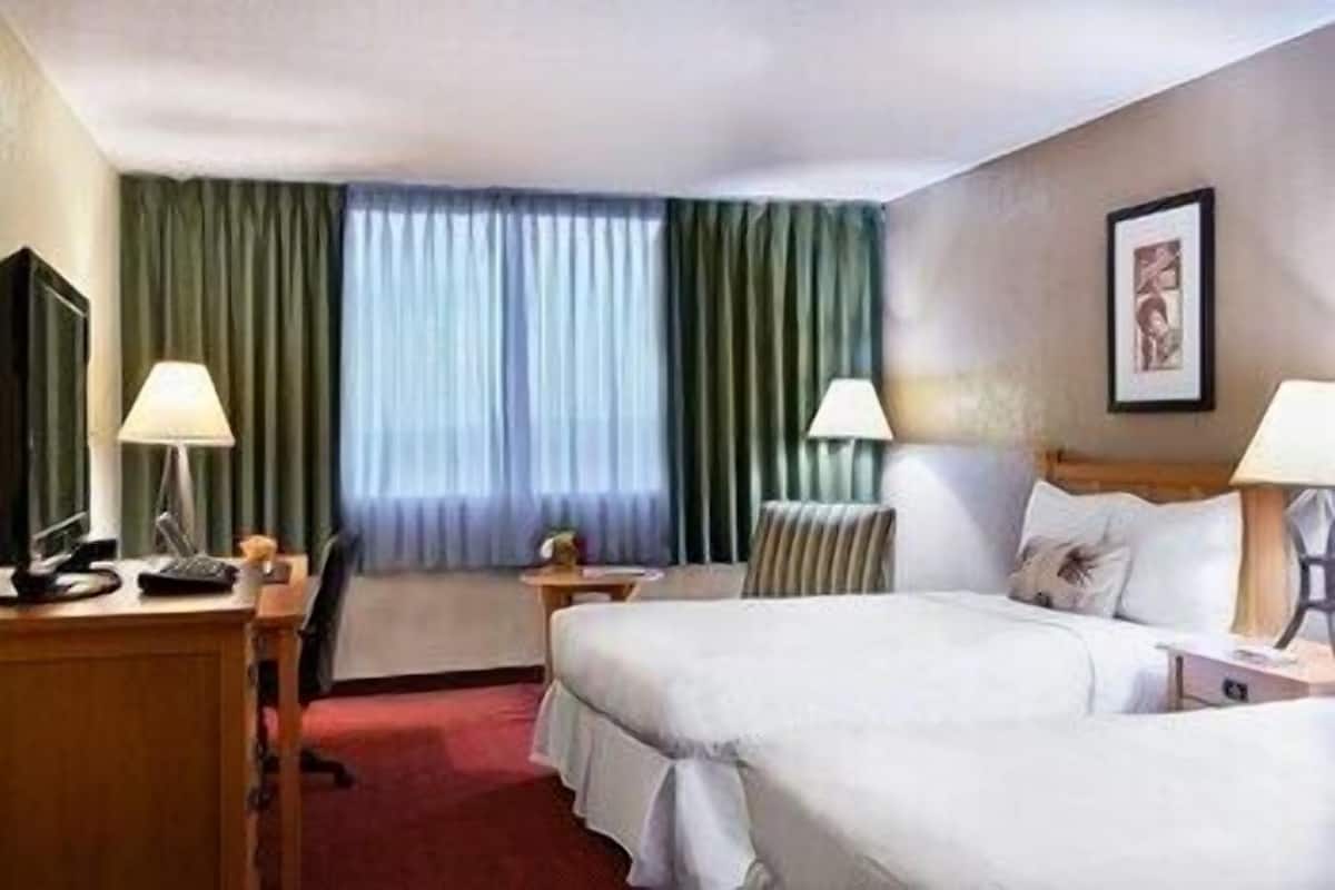 Ein Hotelzimmer mit zwei Betten, einem Schreibtisch, einem Stuhl, einem Fernseher und einem Fenster mit Vorhängen.