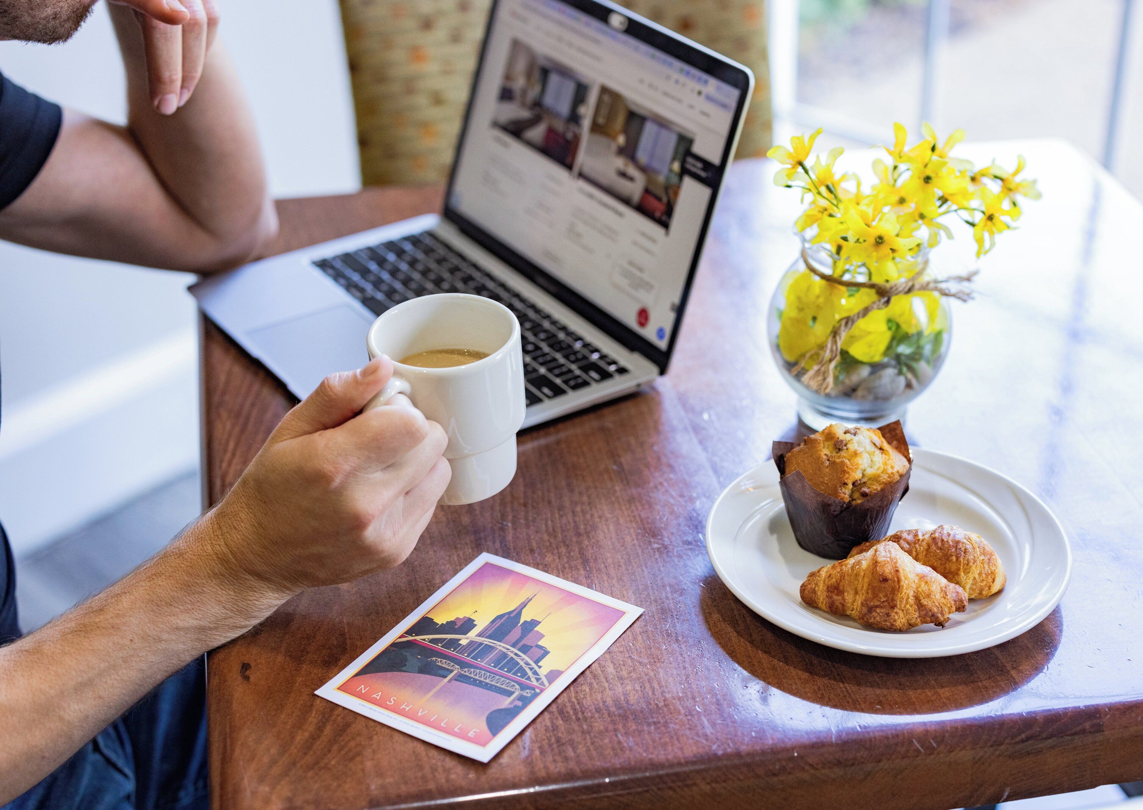 A person holding a white mug, a laptop, a plate with pastries, a vase with yellow flowers, and a postcard on a wooden table.