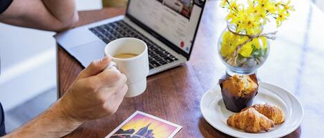 A person holding a white mug, a laptop, a plate with pastries, a vase with yellow flowers, and a postcard on a wooden table.