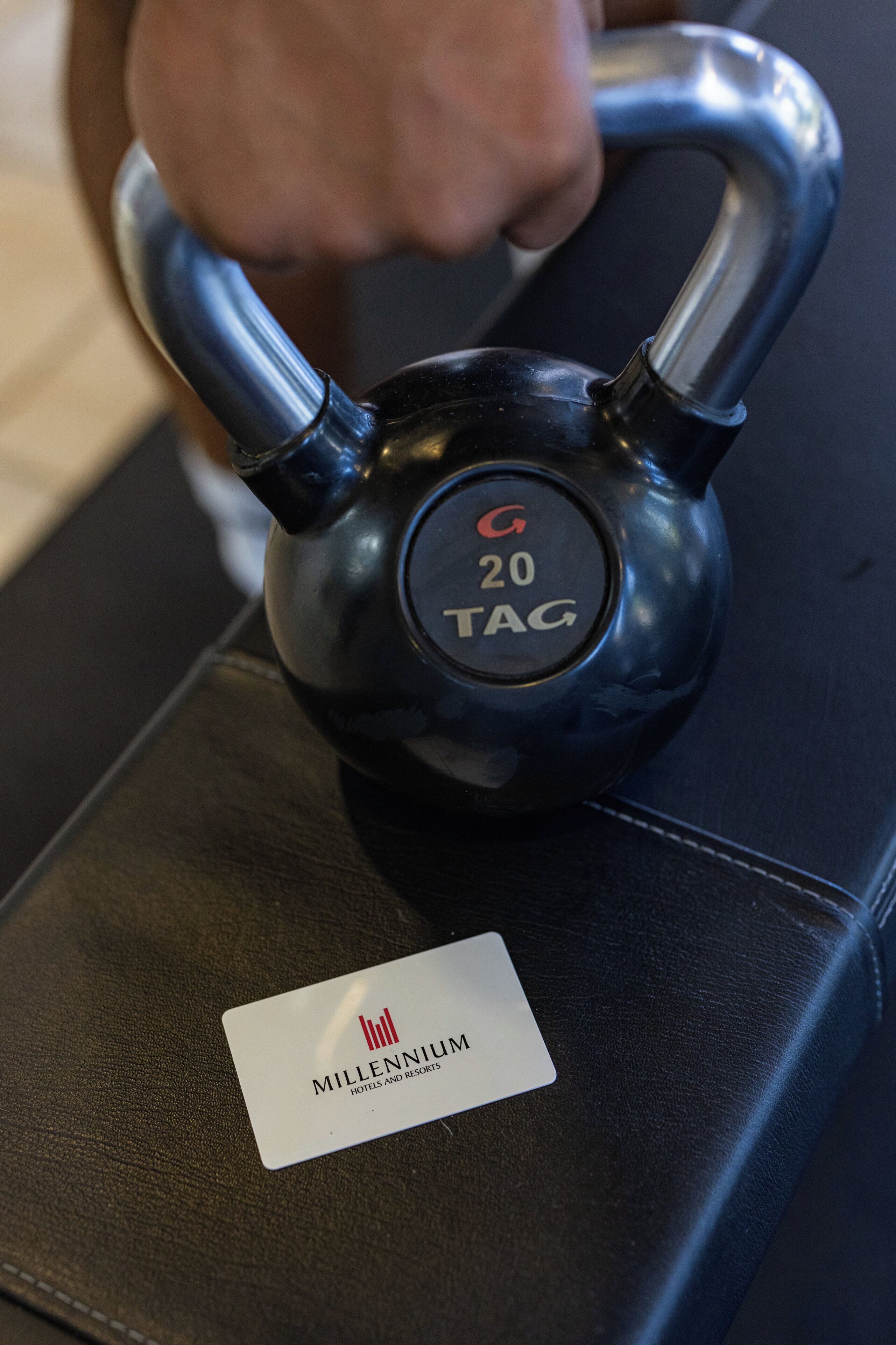 Um kettlebell preto com a inscrição '20 TAG' e o logotipo 'G', apoiado em uma superfície de couro preto com um cartão 'Millennium Hotels and Resorts'.