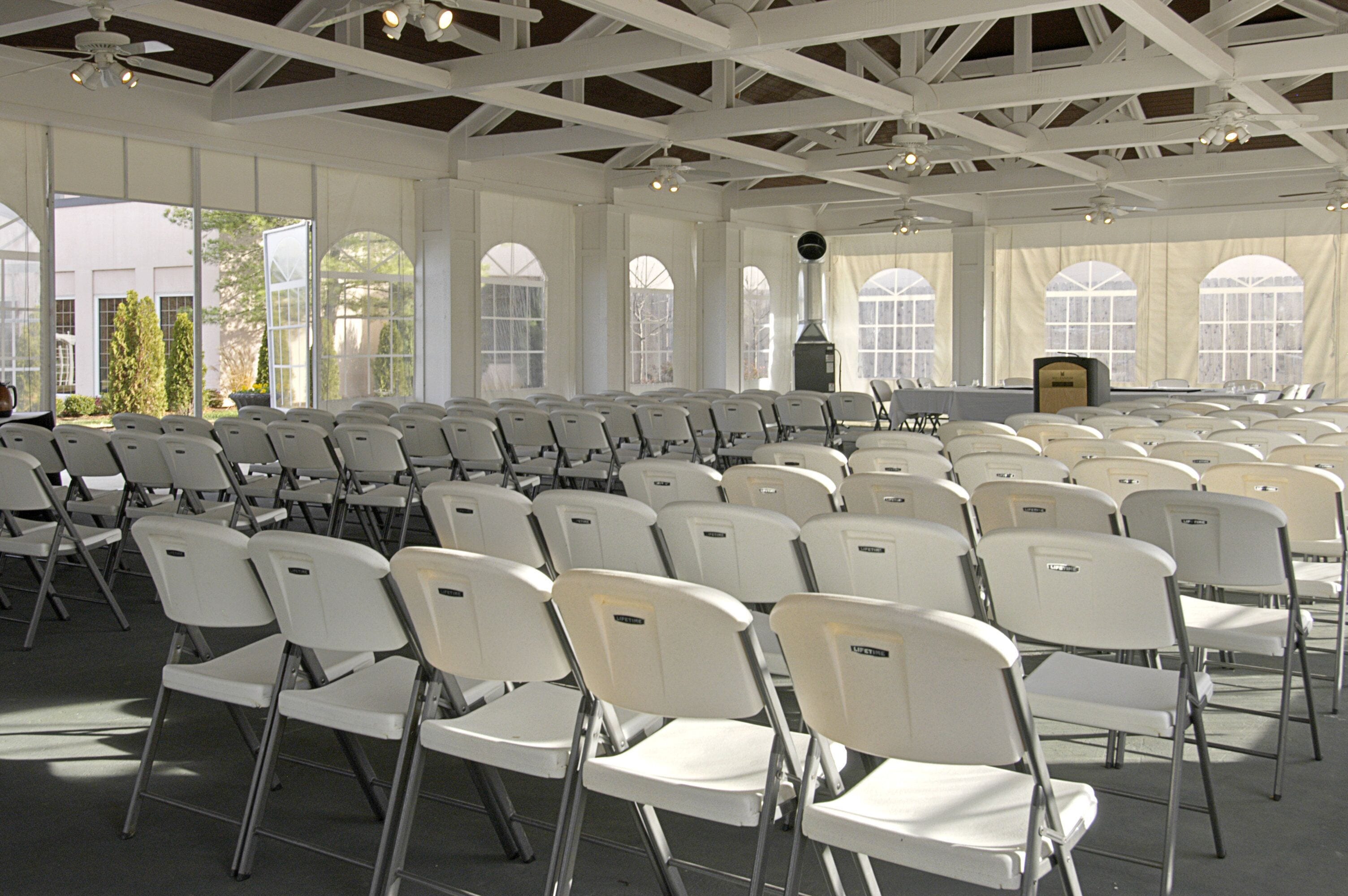 A large indoor space with rows of white folding chairs, a stage with a podium, and large windows allowing natural light.
