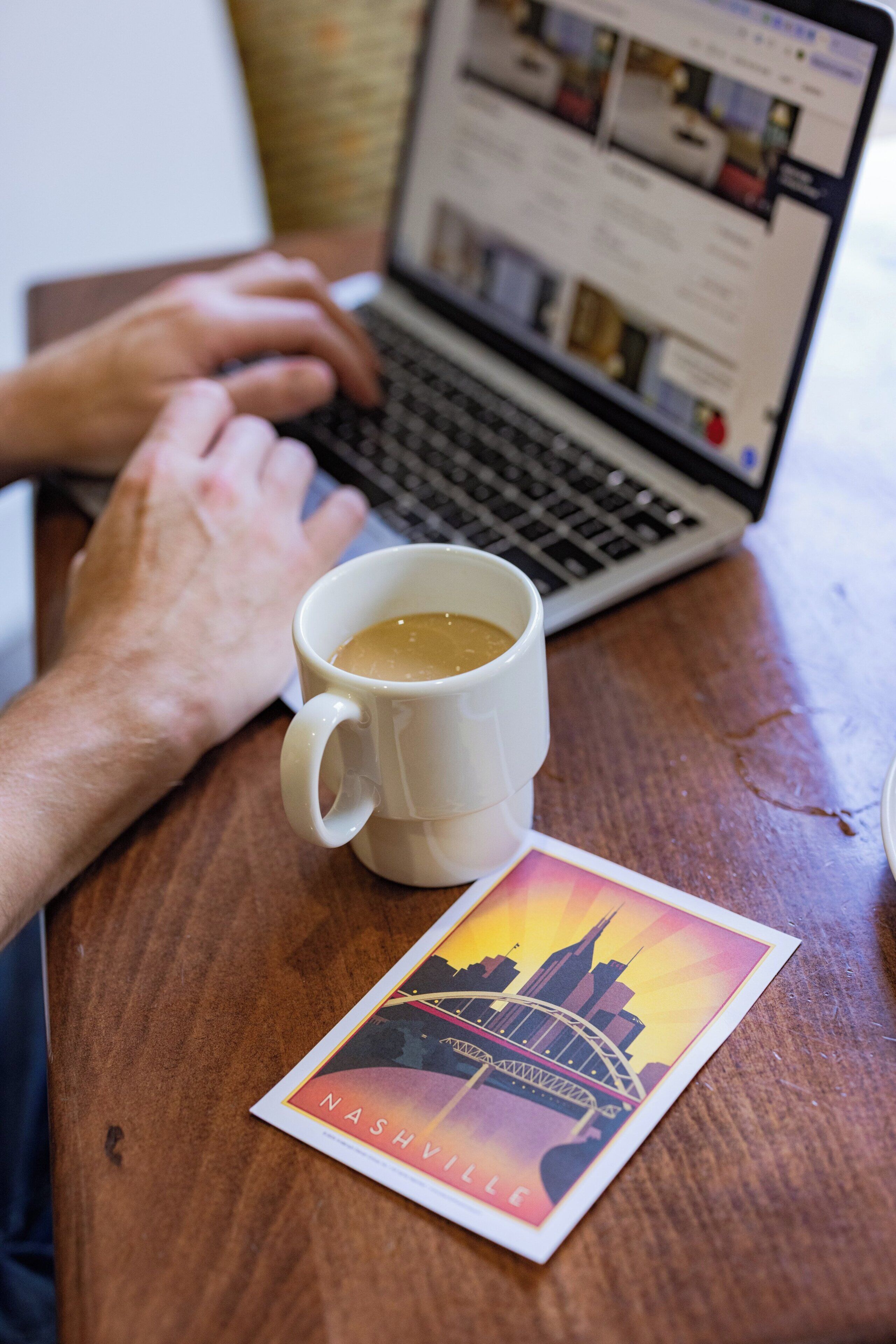 A laptop on a wooden table with a white mug and a postcard of Nashville.