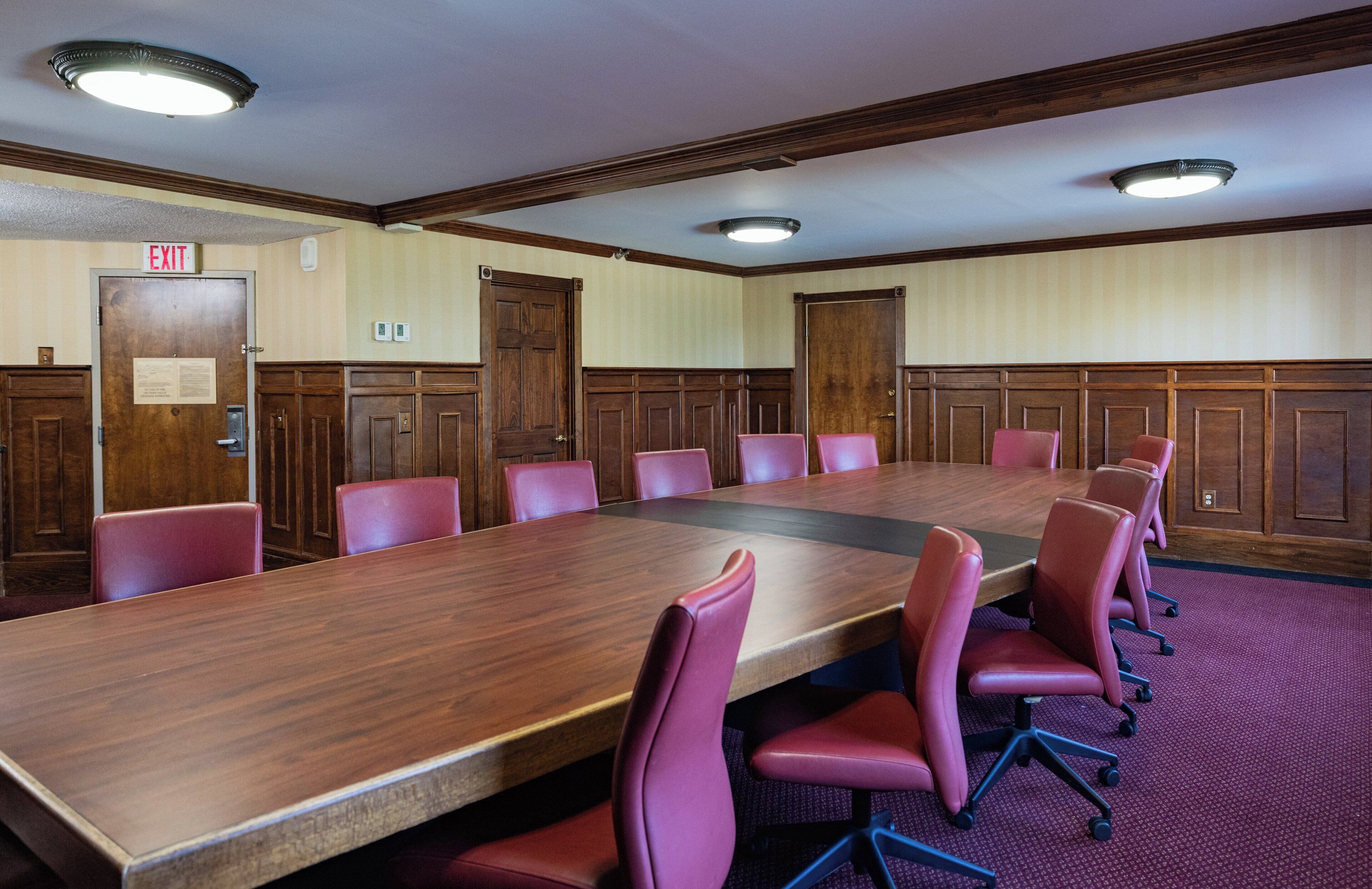 A conference room with a long table, multiple chairs, wooden paneling, and an exit sign.