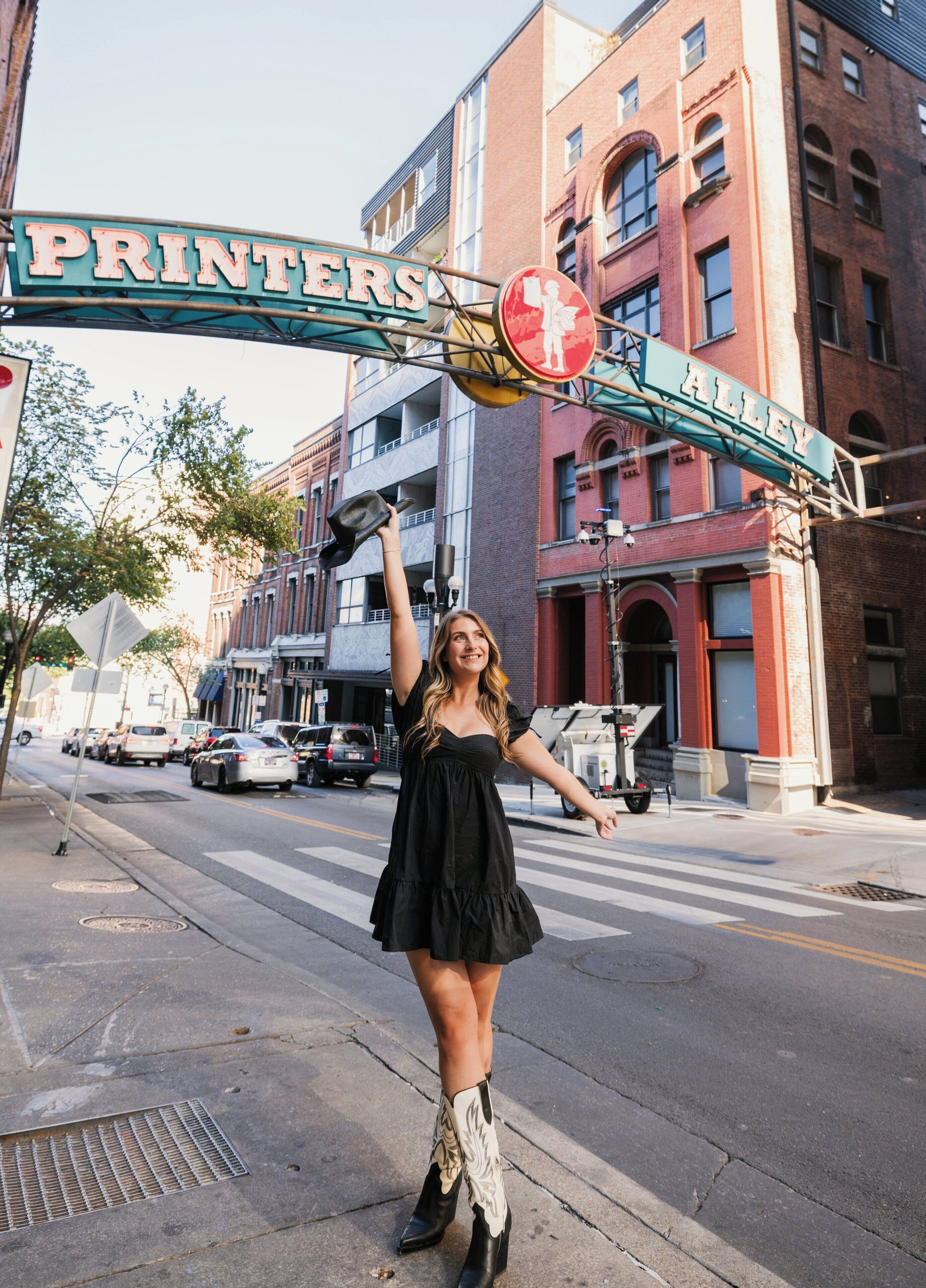 A street scene with a sign reading 'PRINTERS ALLEY' and a woman in a black dress and cowboy boots.