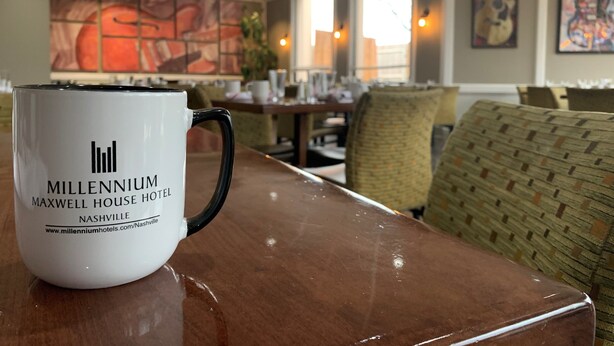 A white mug with "Millennium Maxwell House Hotel Nashville" printed on it, placed on a wooden table in a hotel lobby with wicker chairs and a large abstract painting on the wall. - Millennium Hotel Maxwell House Nashville (Nashville)