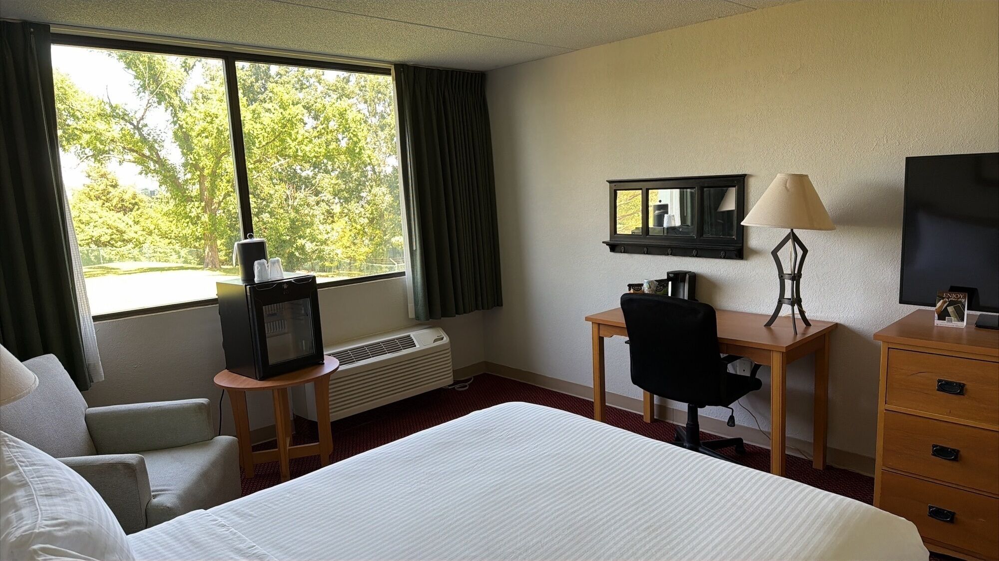 Una habitación de hotel con cama, escritorio con silla, televisor, calefacción y ventana con cortinas.