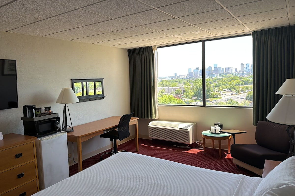 Ein Hotelzimmer mit Bett, Schreibtisch, Stuhl, Fernseher und einem Fenster mit Blick auf die Stadt.