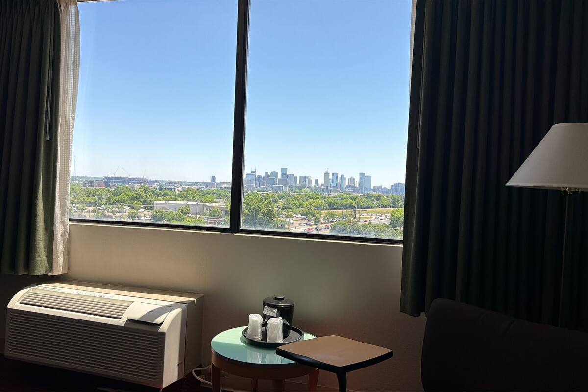 Ein Hotelzimmer mit großem Fenster, Klimaanlage, Tisch mit Lampe und Blick auf einen Stadt-Horizont.