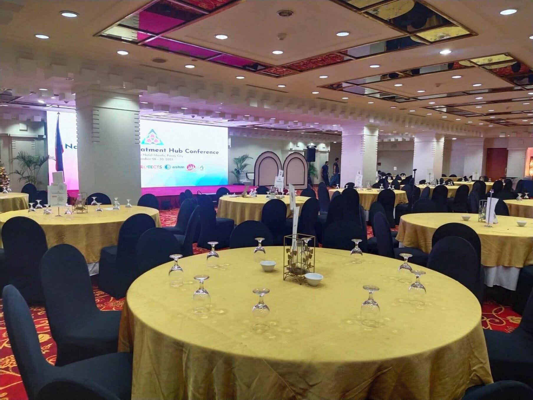 Un salón de conferencias con mesas redondas, sillas negras y un escenario con una pantalla de presentación.