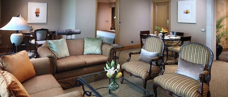 A hotel room with a living area featuring a sofa, armchairs, a glass coffee table, and a dining table with chairs.