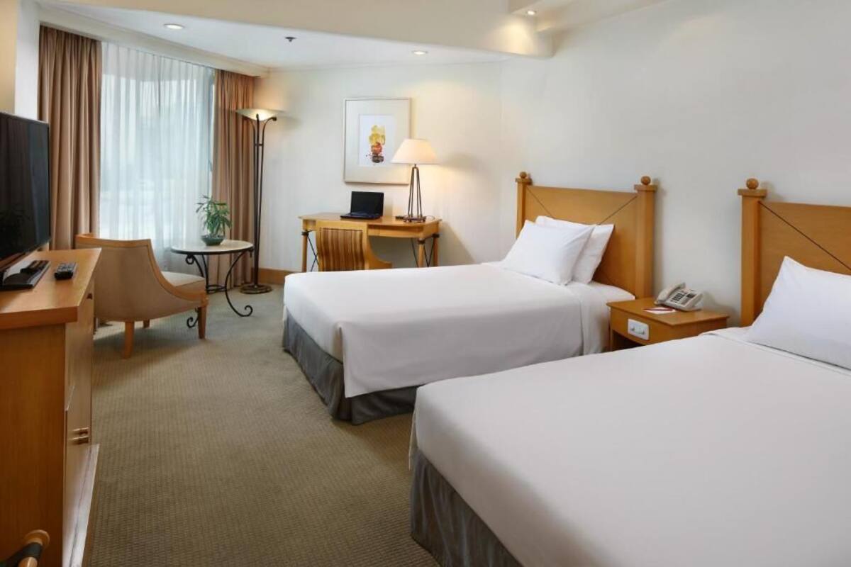 A hotel room with two single beds, a desk with a computer, a chair, a TV, and a lamp.