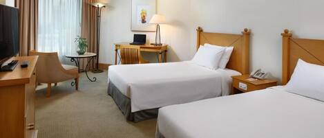 Quarto de hotel com duas camas de solteiro, uma escrivaninha com computador, uma cadeira, televisão e luminária.