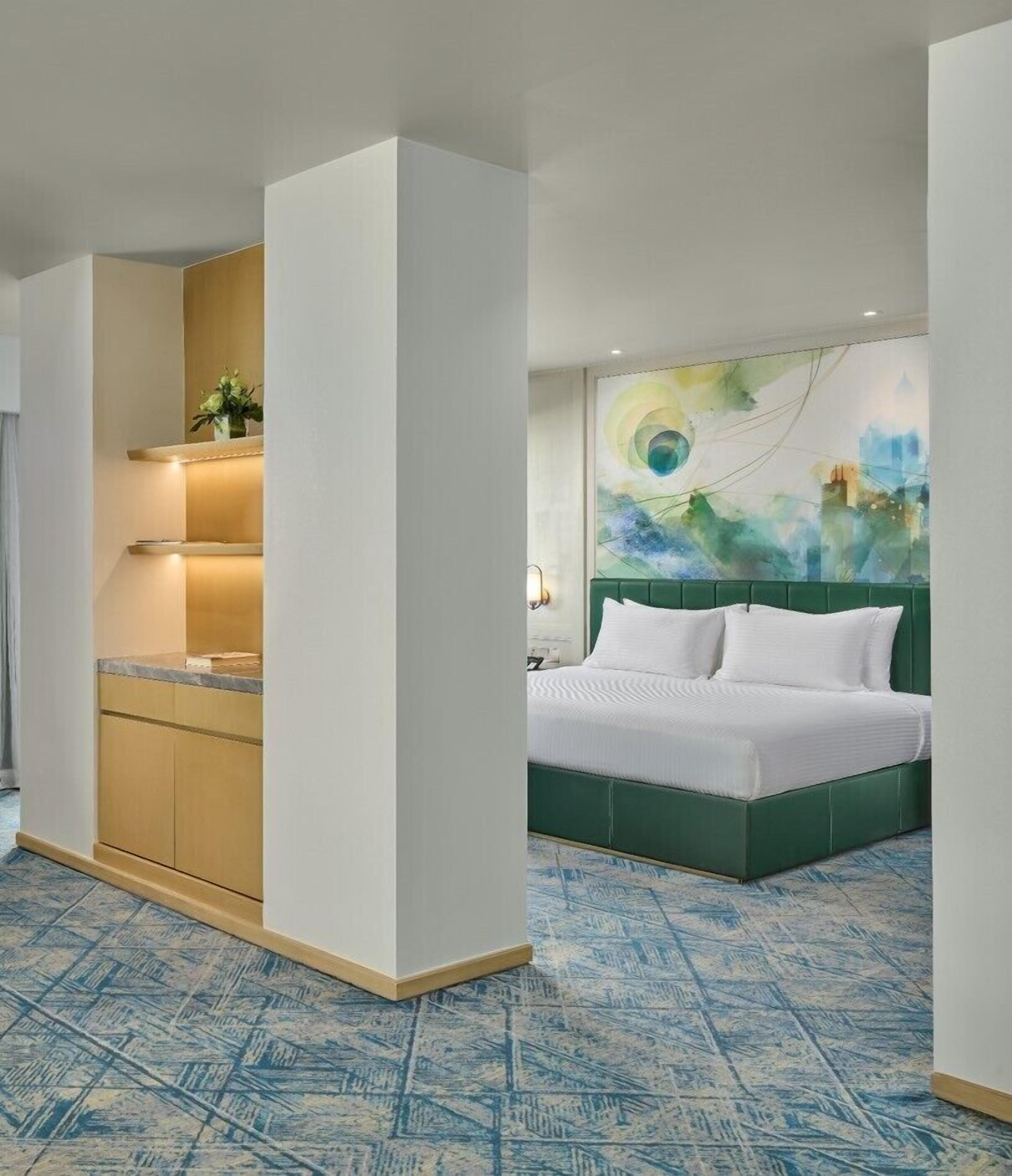 A hotel room with a green bed, a white headboard, a blue patterned floor, a kitchen area with a countertop and shelves, and a wall with a colorful abstract painting.