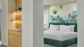 Habitación de hotel con cama verde, cabecera blanca, suelo de diseño azul, área de cocina con encimera y estantes, y pared con un cuadro abstracto de colores.