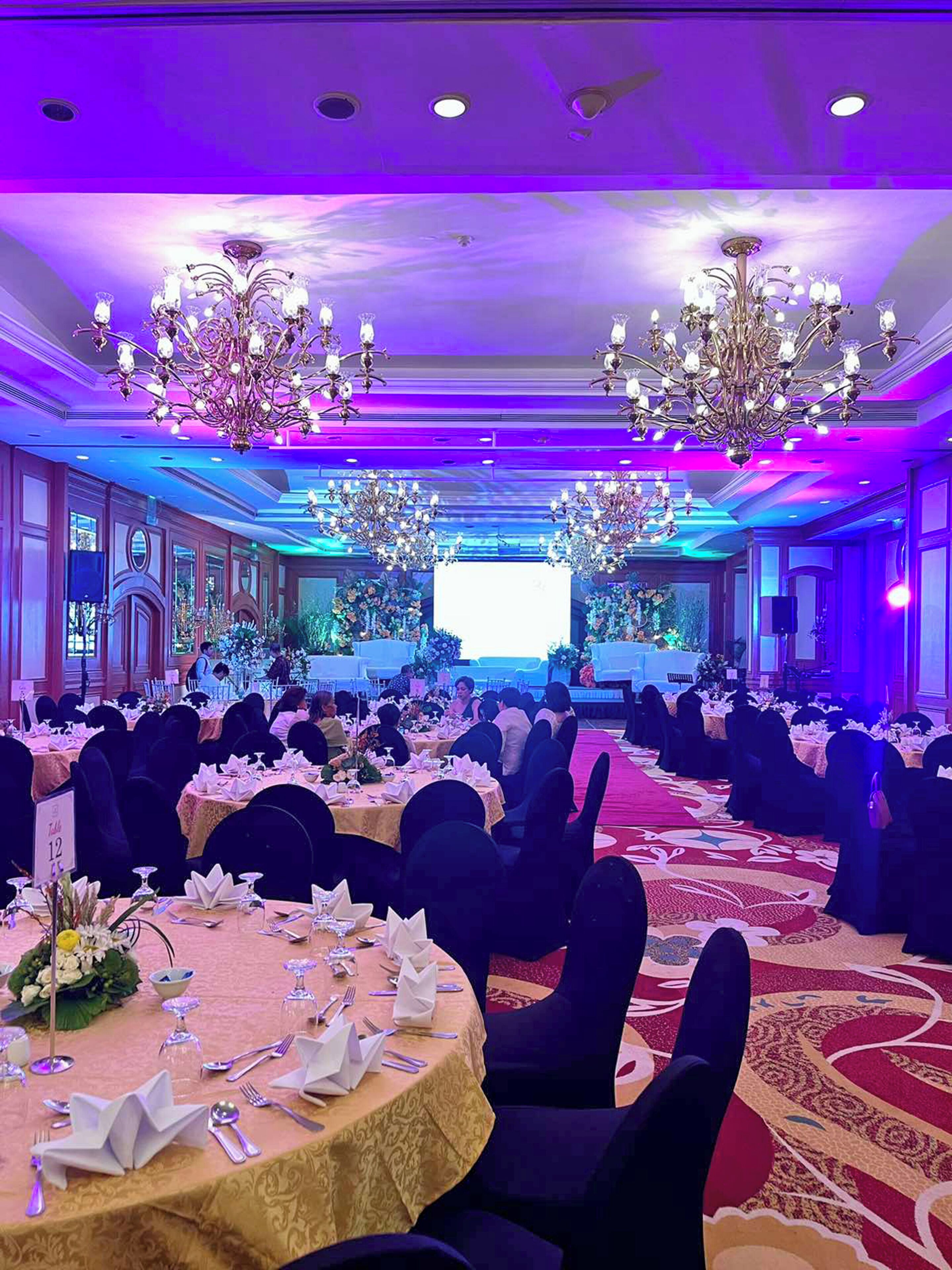 A banquet hall with round tables, black chairs, and chandeliers.