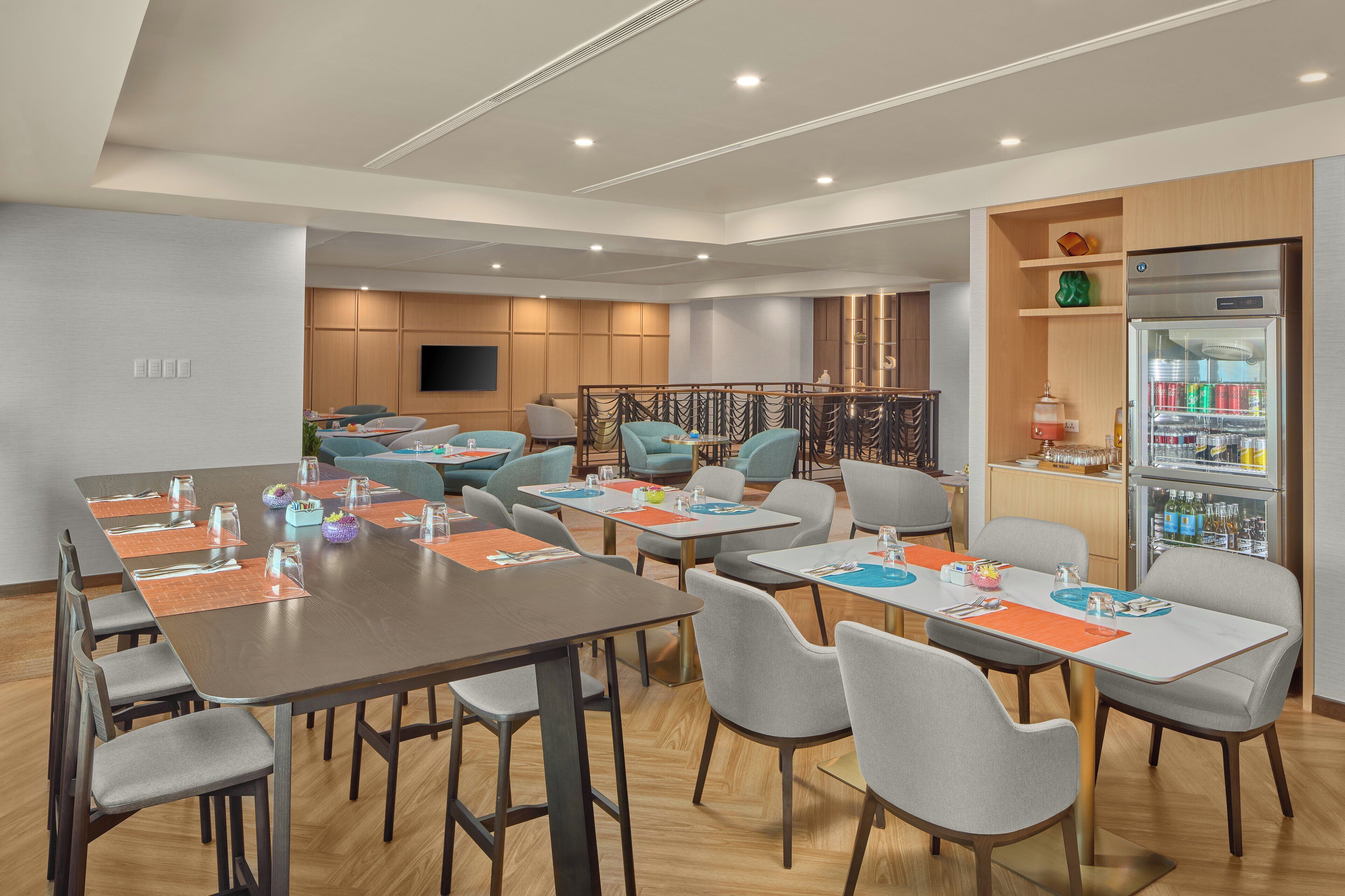 A modern dining area with multiple tables and chairs, a kitchen area with a refrigerator and shelves, and a seating area with sofas and a TV.