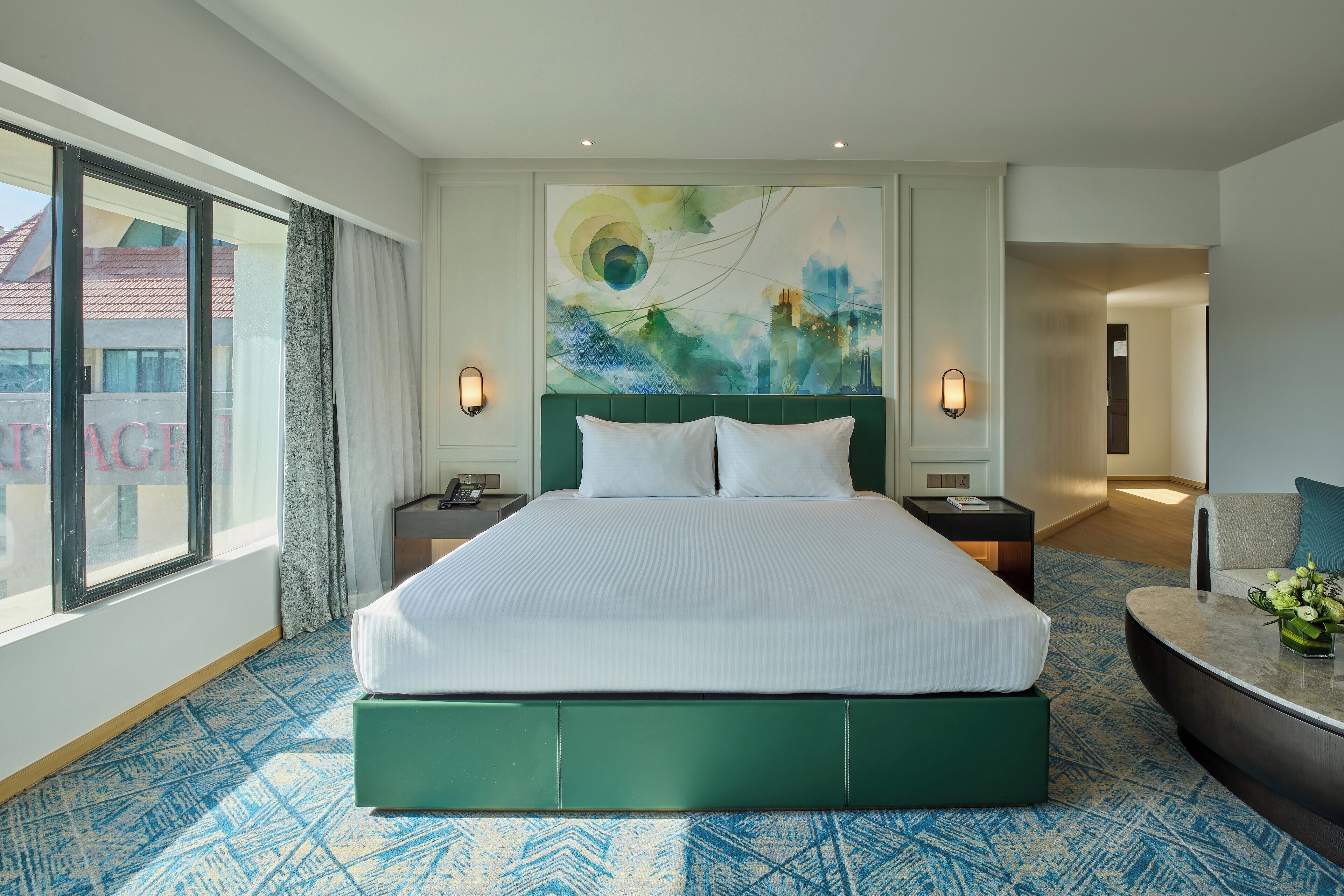 A hotel room with a large bed, two bedside tables, a nightstand, a desk with a chair, a large window, and a wall painting.