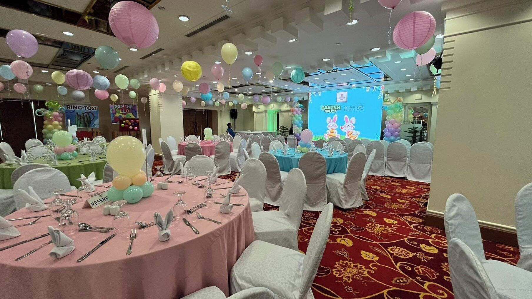 Une salle de banquet avec des tables rondes dressées pour un événement formel, décorée de ballons et d’une banderole pour une célébration de Pâques.