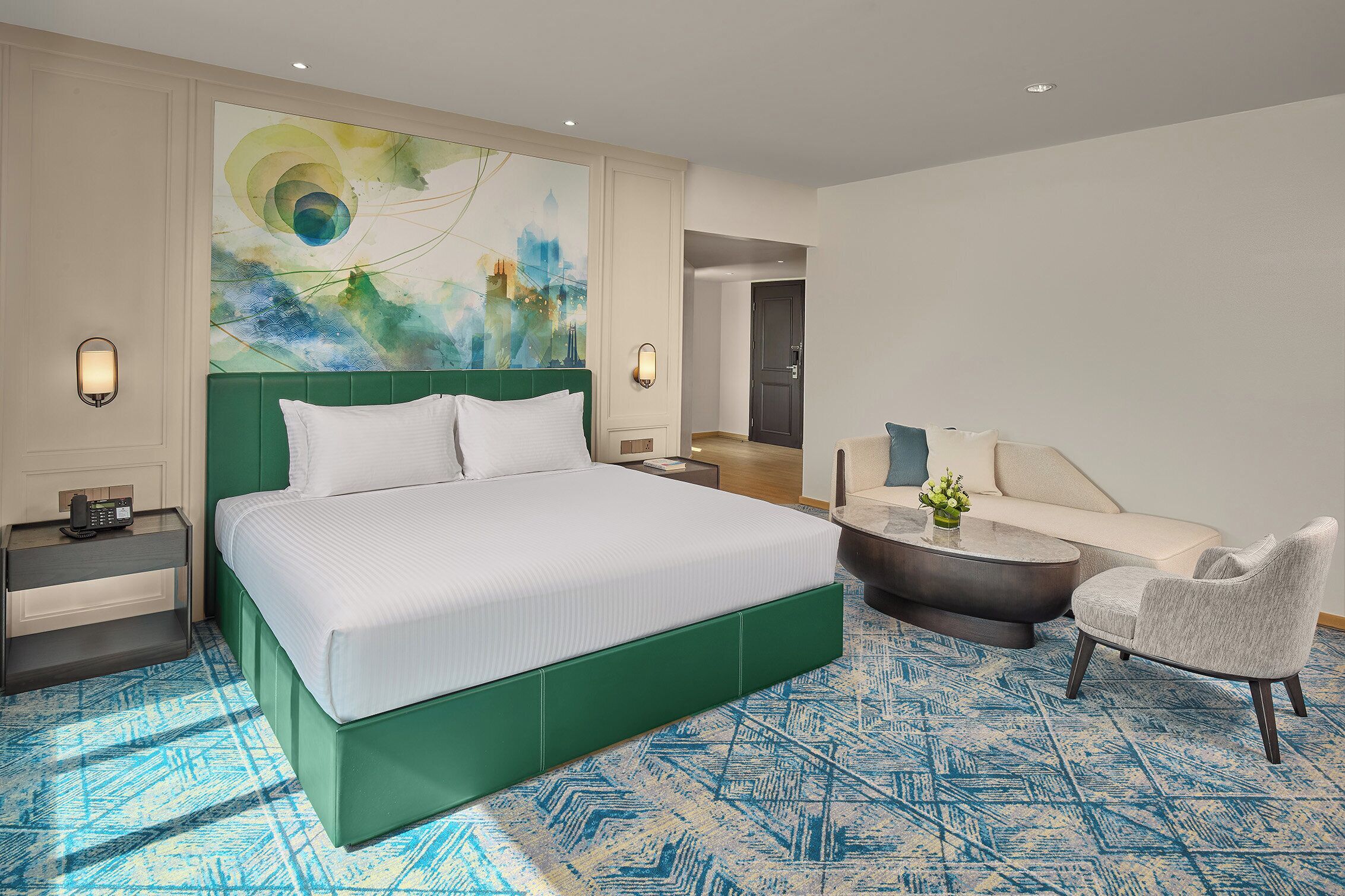 A modern hotel room with a large bed, a green headboard, a sofa, a chair, and a coffee table.