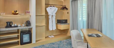 Habitación de hotel con un escritorio de madera, una silla, un refrigerador pequeño, una cafetera, un microondas, un cuenco de fruta y una repisa con botellas y vasos.