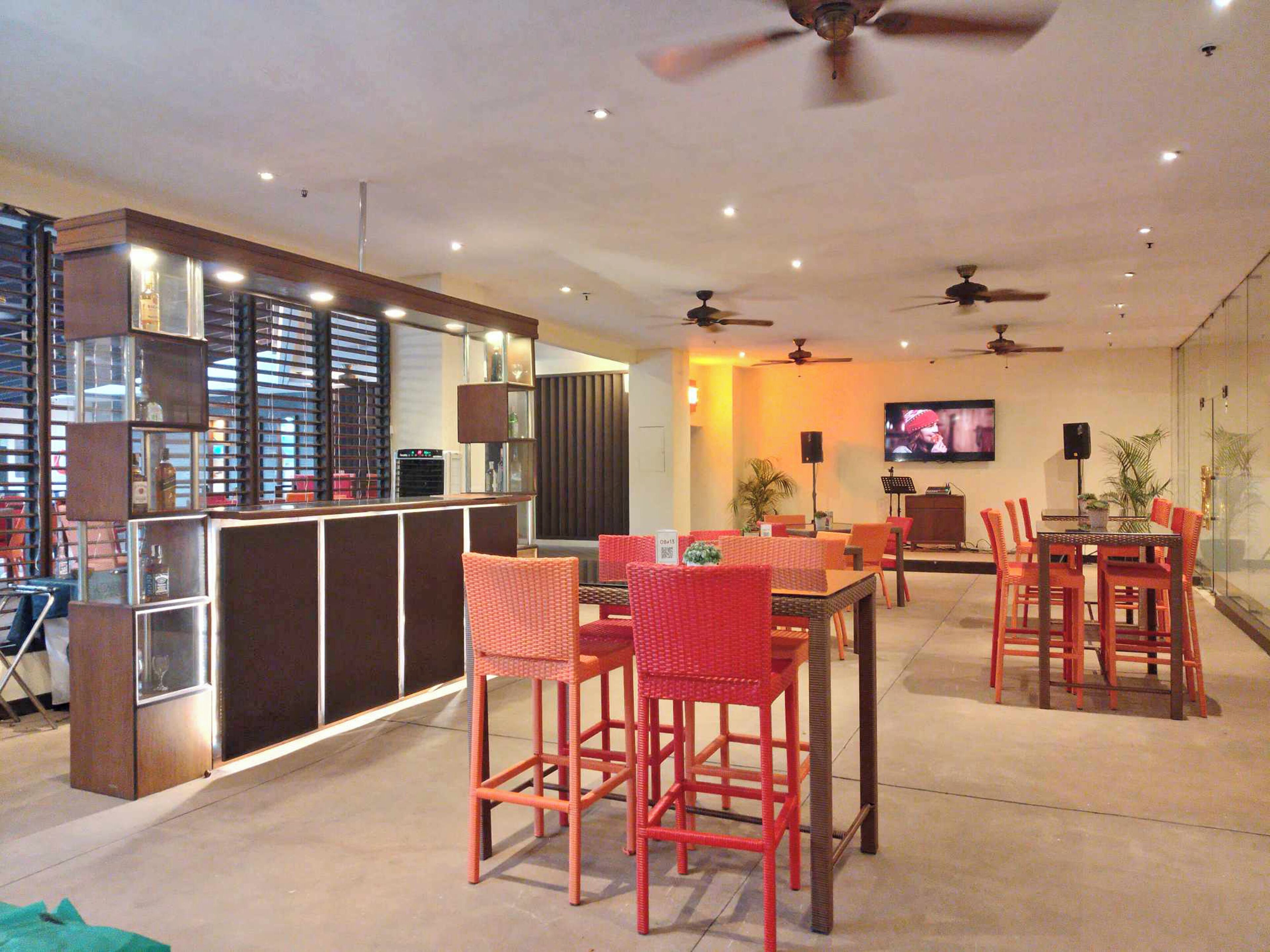 A modern bar area with a counter, seating with red chairs, ceiling fans, and a TV screen.