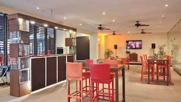 Un área de bar moderna con mostrador, asientos con sillas rojas, ventiladores de techo y una pantalla de televisión.