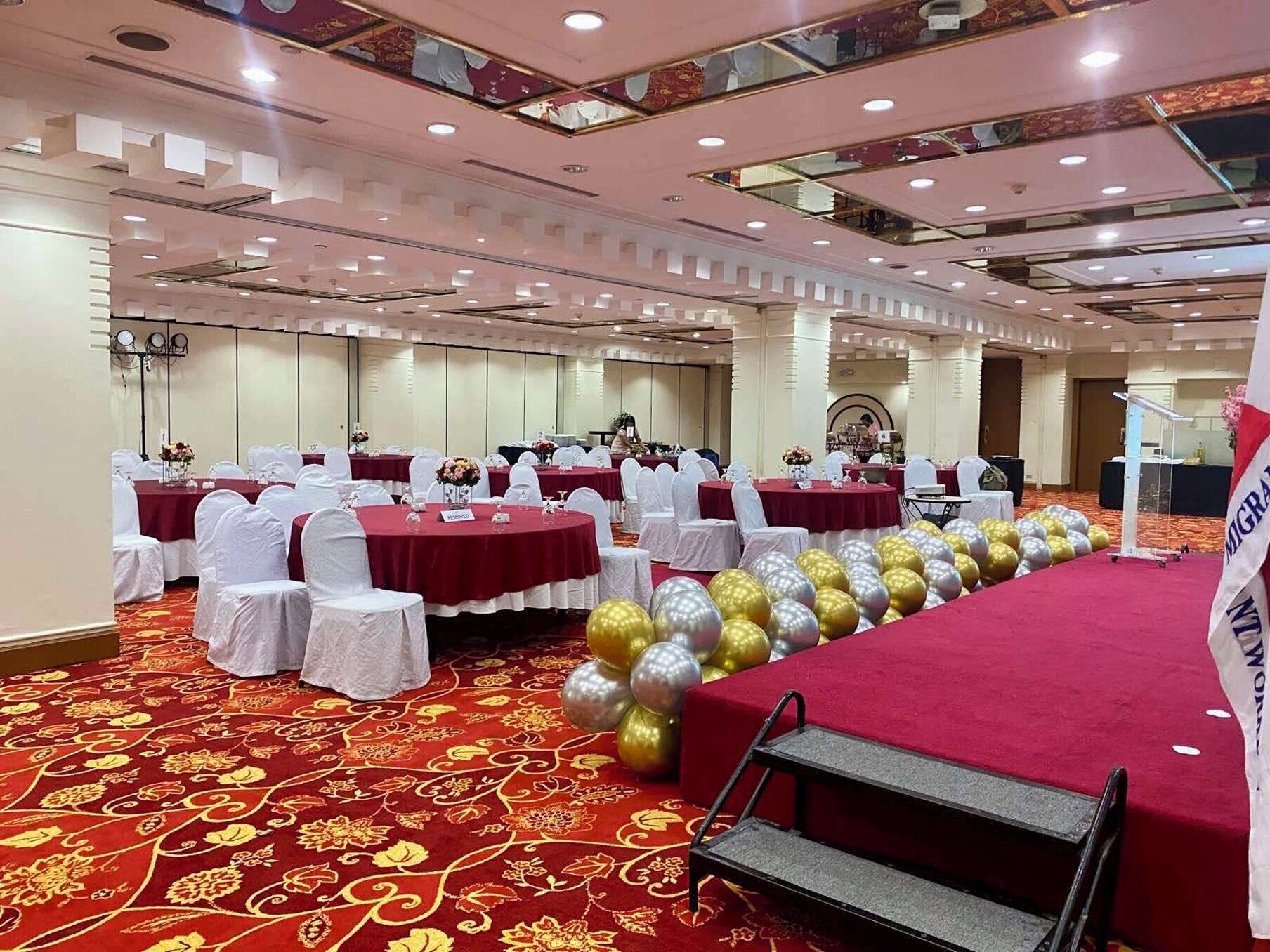 Un salón de banquetes con mesas redondas cubiertas con manteles blancos y caminos de mesa rojos, dispuestas en filas.