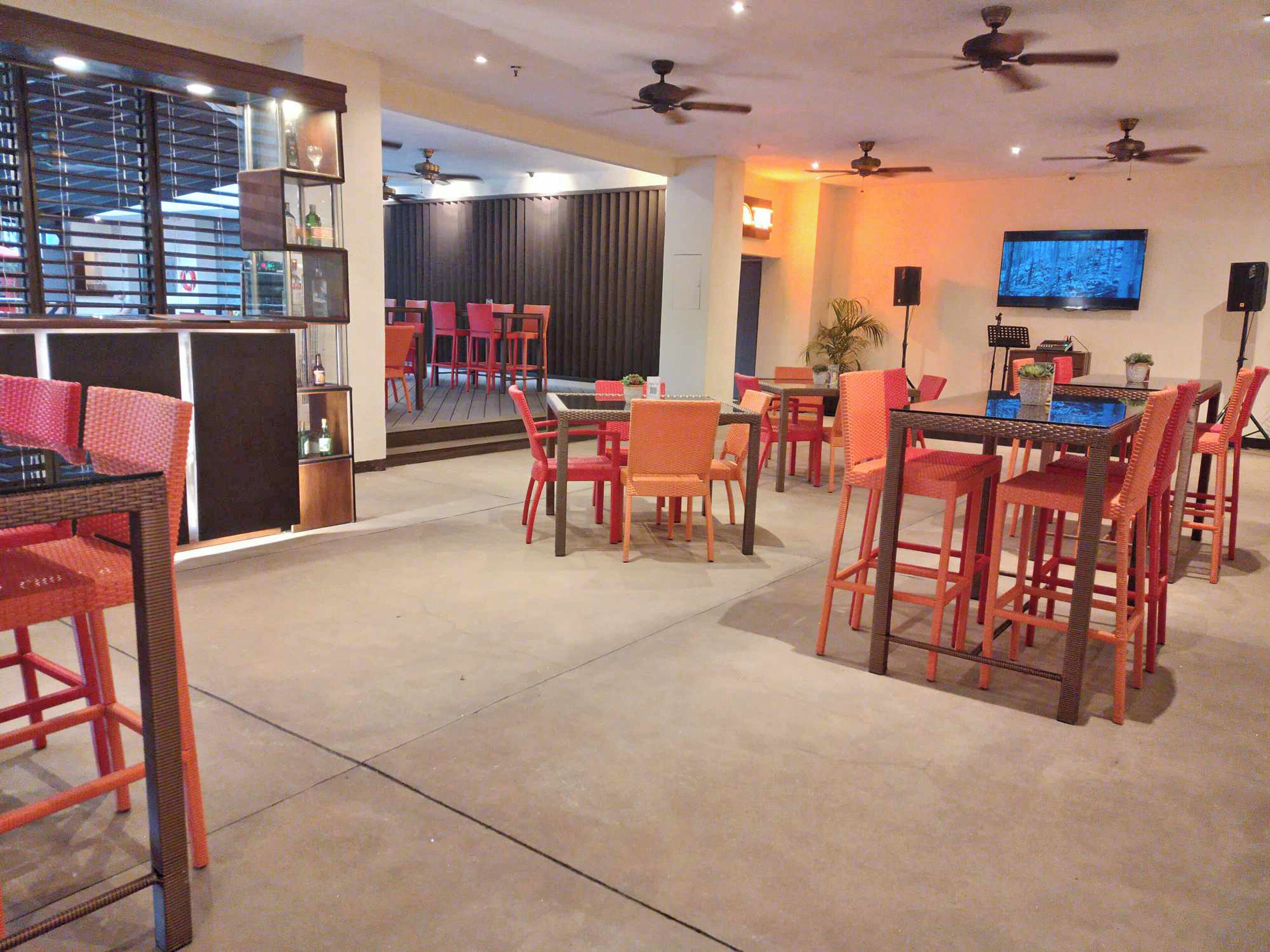 A spacious indoor area with multiple seating arrangements, including a bar area with a counter and chairs, and a dining area with tables and chairs. There are ceiling fans and a television mounted on the wall.